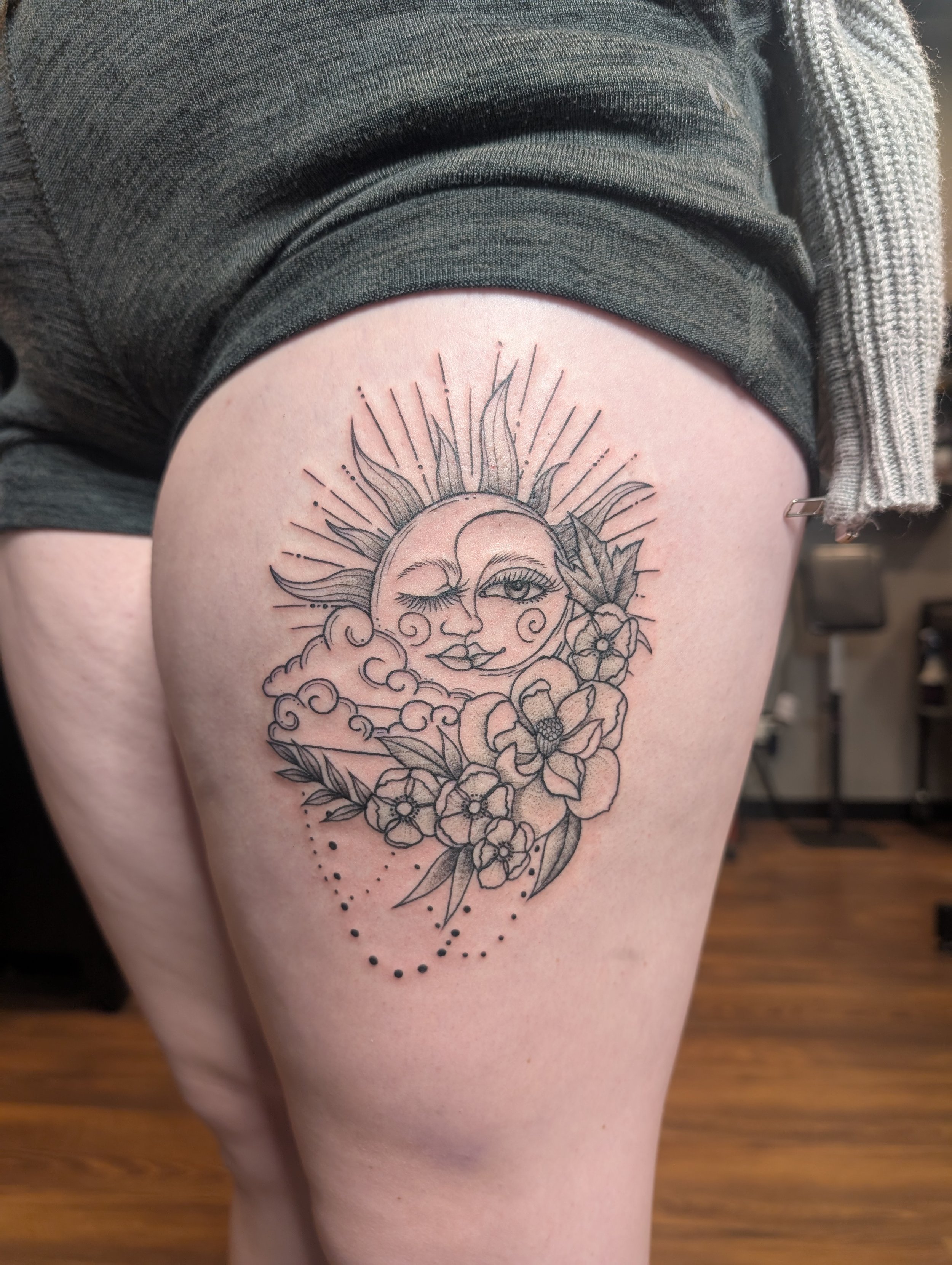 Tattoo of a smiling sun with a face, winking eye, surrounded by flowers and leaves, with dotted accent details.