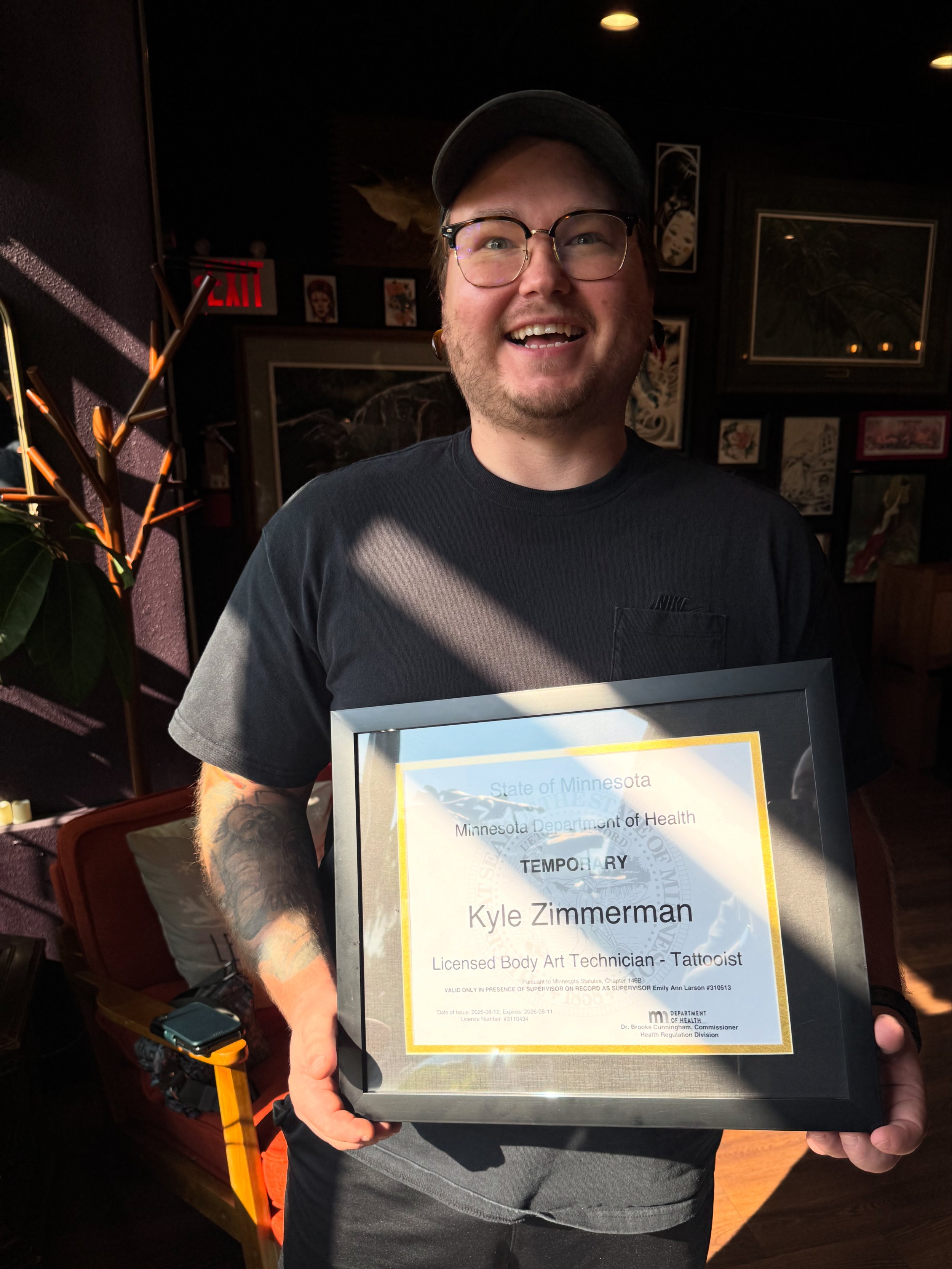 Man holding a framed certificate, smiling, in a dimly lit room with artwork and plants.