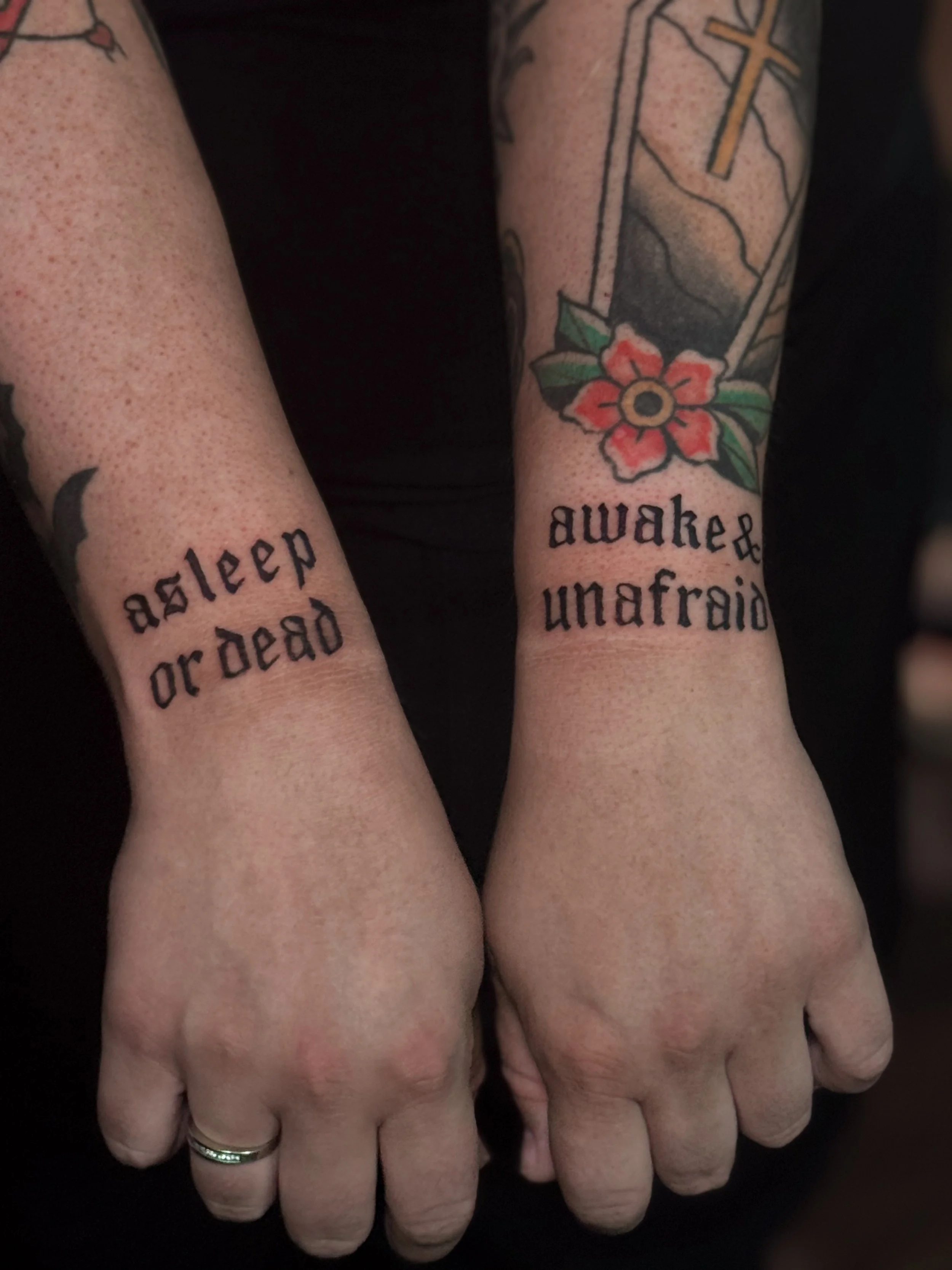 Two hands with tattoos. The left hand has the words 'asleep and dead' tattooed on the wrist, while the right hand has a tattoo of a cross, mountain landscape, and a red flower with green leaves. The right hand also has the words 'awake & unafraid' ta