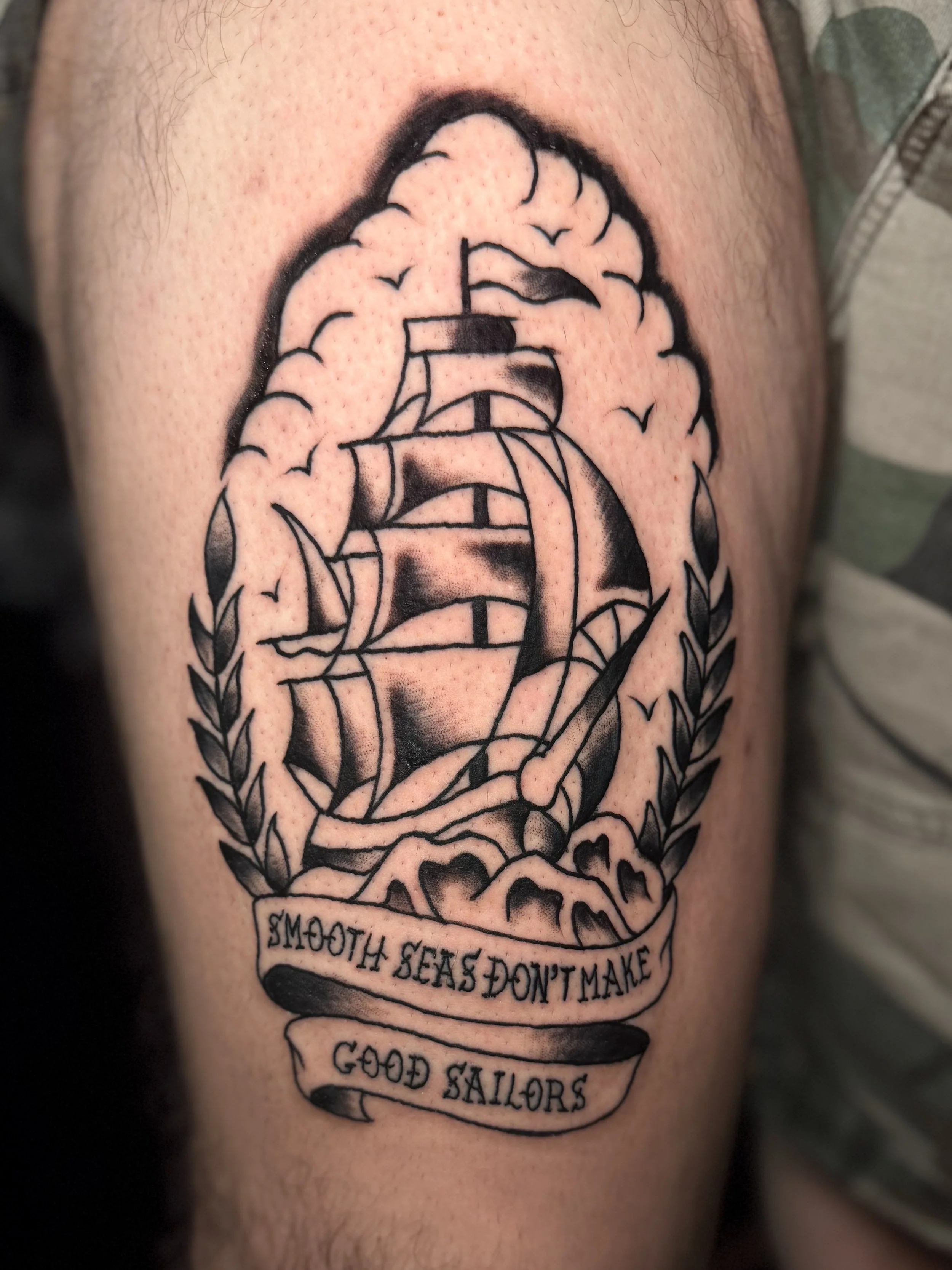 A tattoo of a sailing ship with waves and birds above it on someone's shoulder, with text saying 'Seafood & Smoke, No Seas Don't Make, Good Sailors'.
