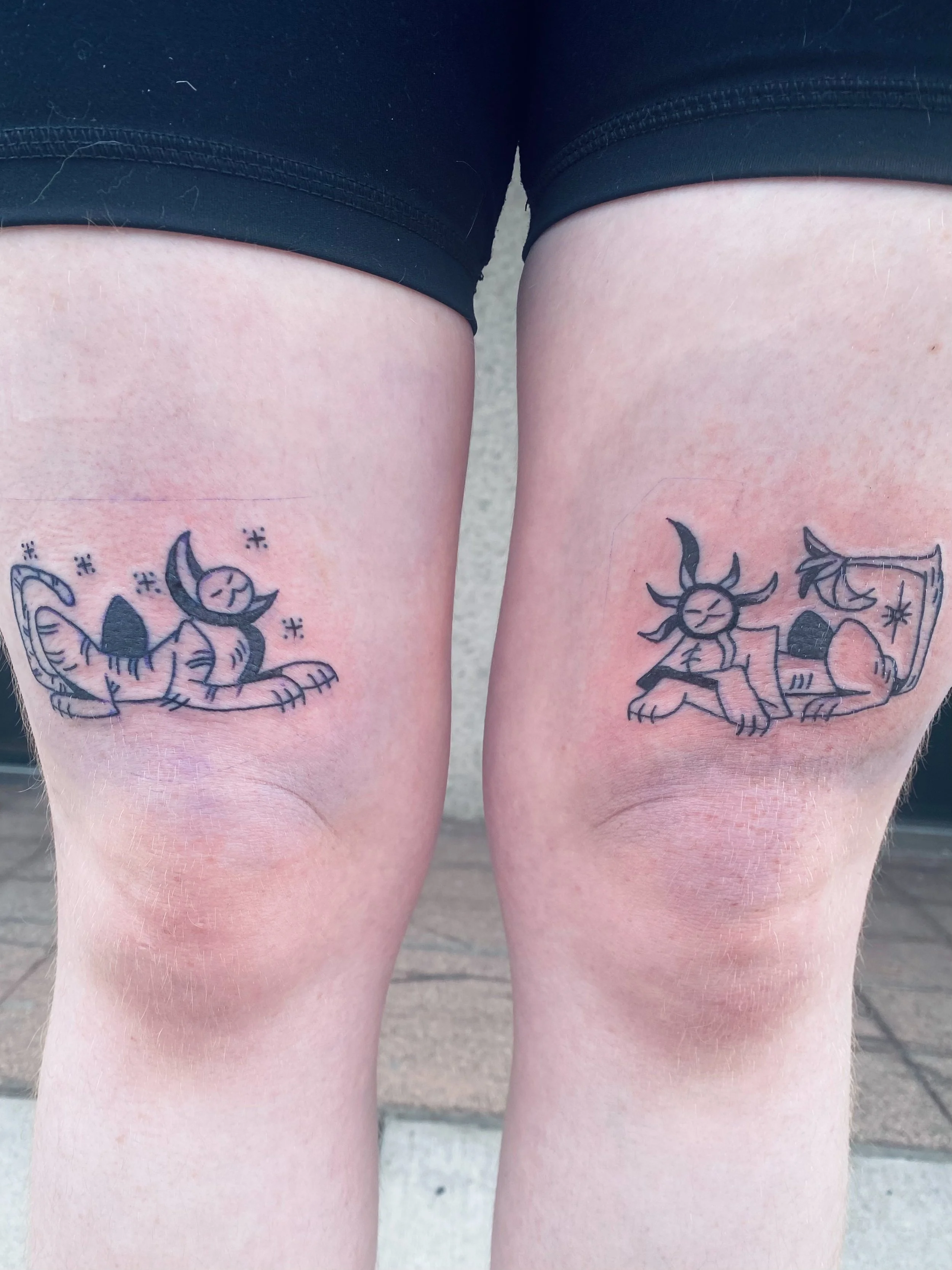 Close-up of two person's upper thighs with tattoos, wearing black shorts, standing outdoors.