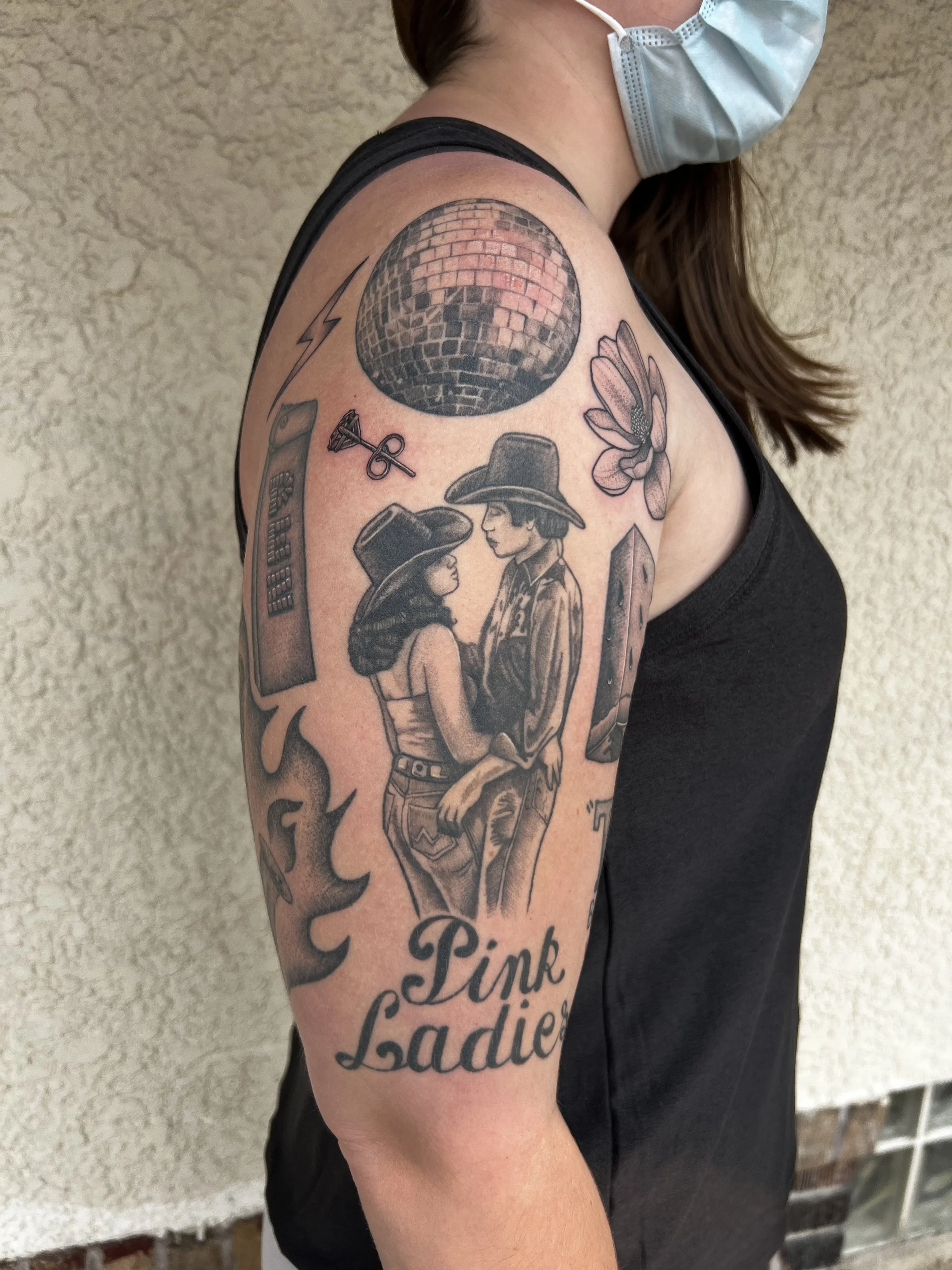 A woman with a shoulder tattoo, wearing a face mask and black tank top, standing against a textured beige wall.