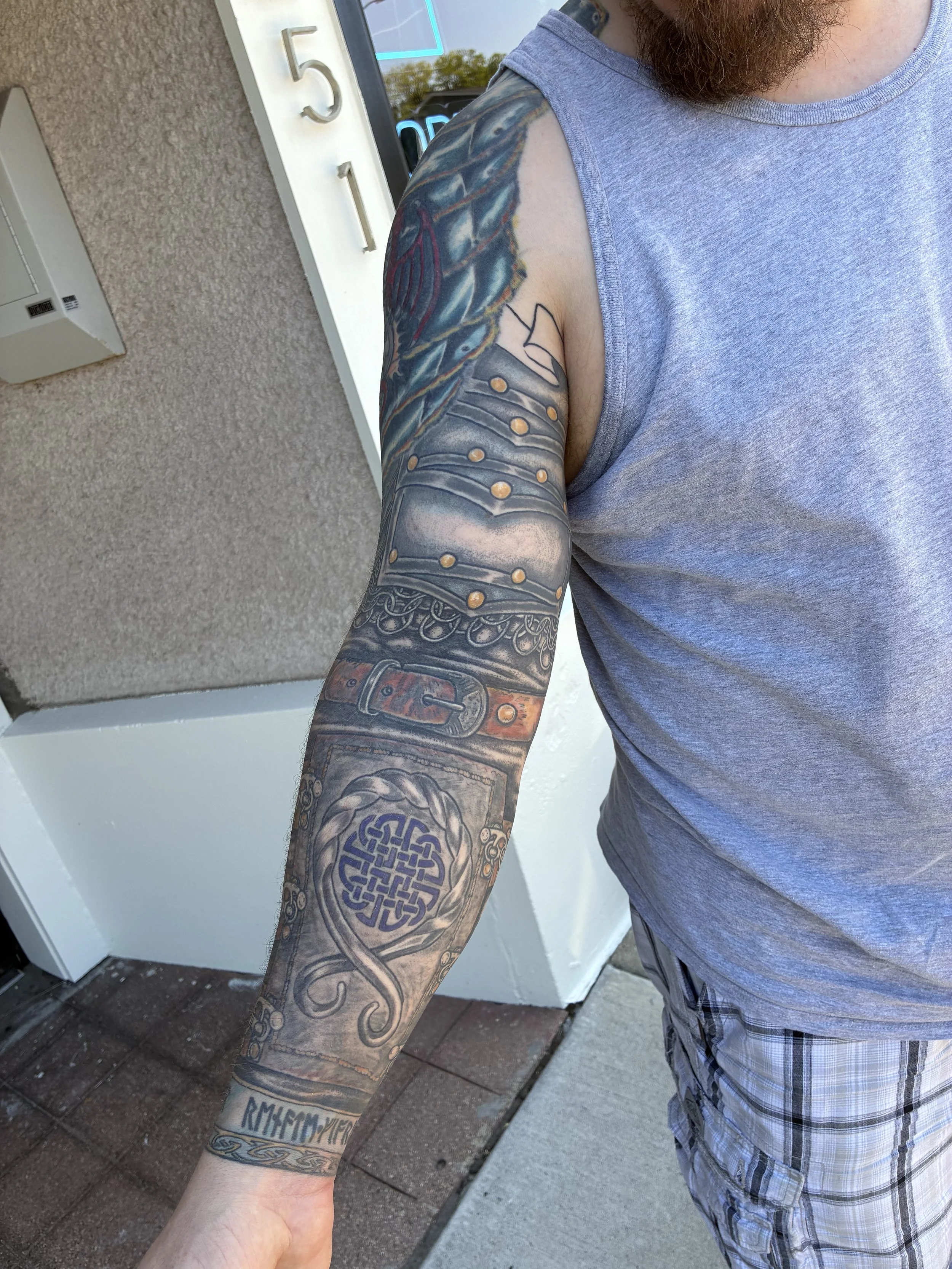 Close-up of a man's arm with tattoos, wearing a sleeveless gray shirt, standing outside near a white building with beige textured wall, window, and door.
