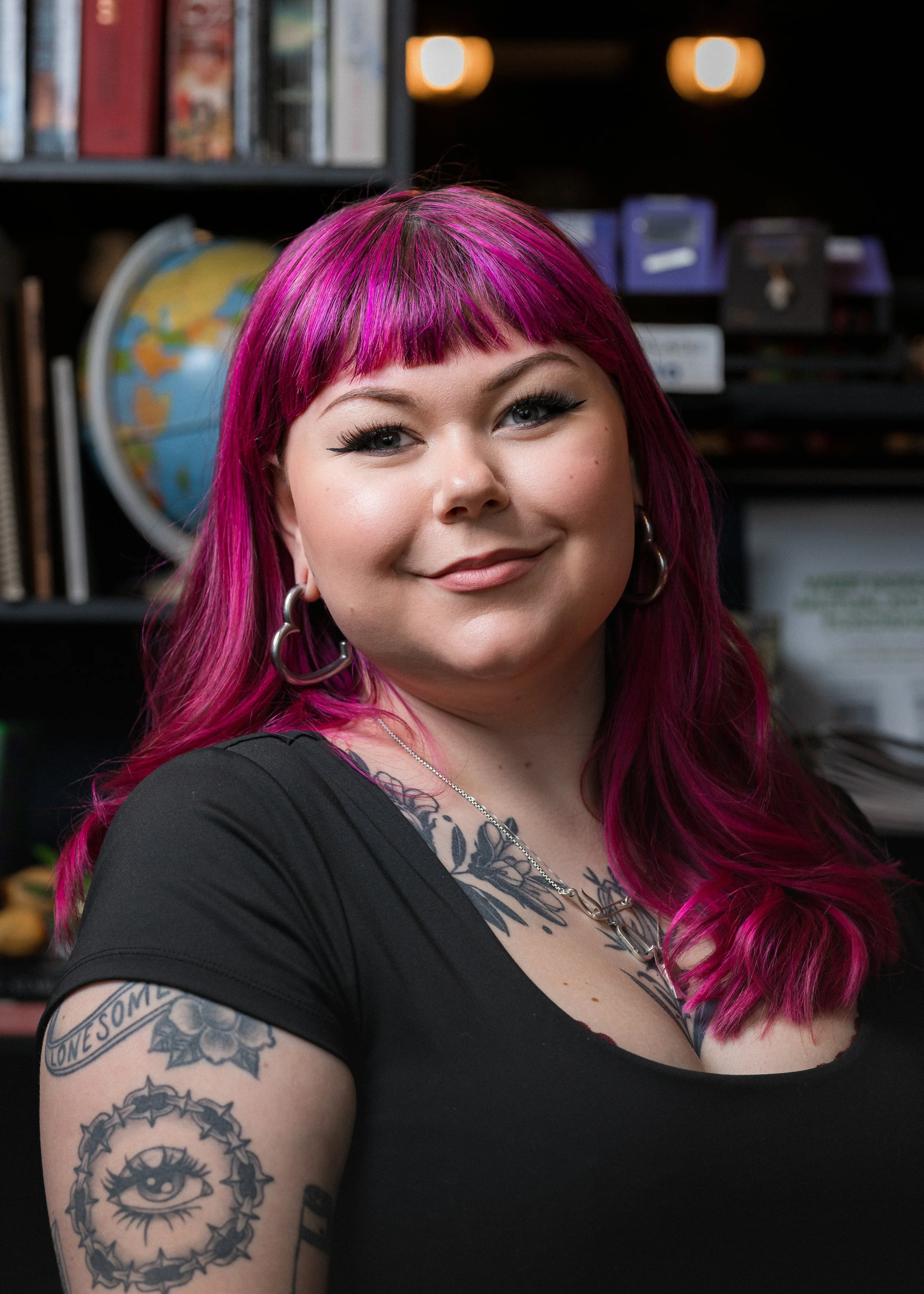 A woman with vibrant pink hair styled with bangs, wearing eyeliner and silver hoop earrings. She has tattoos on her right arm and chest, including an eye within a crescent moon, a banner with the word "awesome," a star, and decorative designs. She is smiling and appears to be in a room with shelves of books, a globe, and storage boxes in the background.