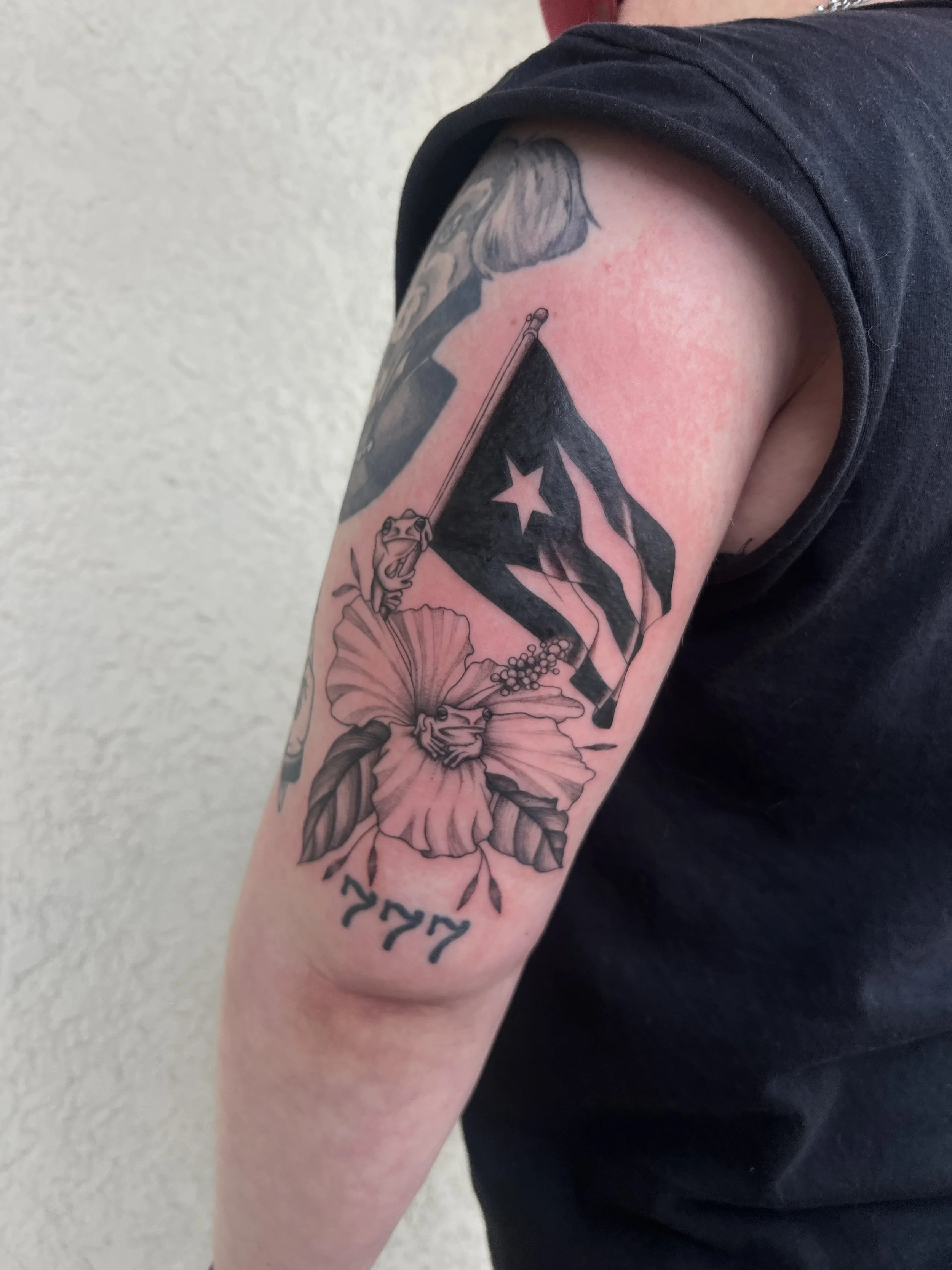 A person's upper arm with a black tattoo of a flag with a star, a key, a lotus flower, and some writing.