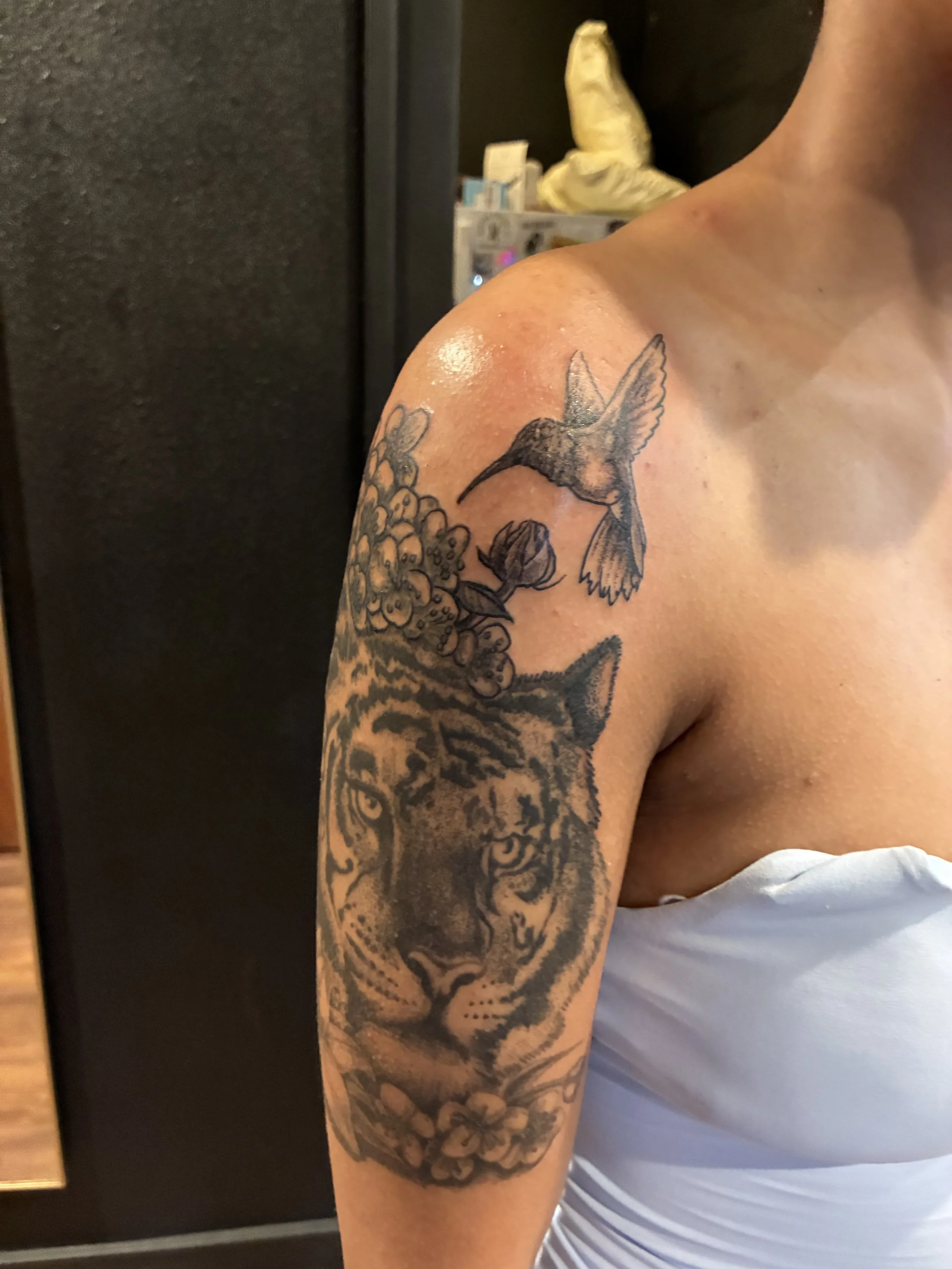 Close-up of a person's upper arm with a black and gray tattoo of a hummingbird and flowers.