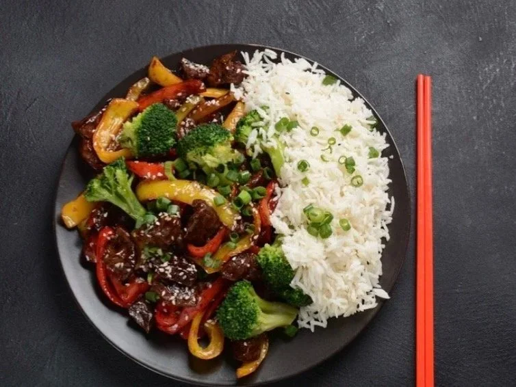 Plate of vegetable stir-fry with rice and red chopsticks on a dark table.