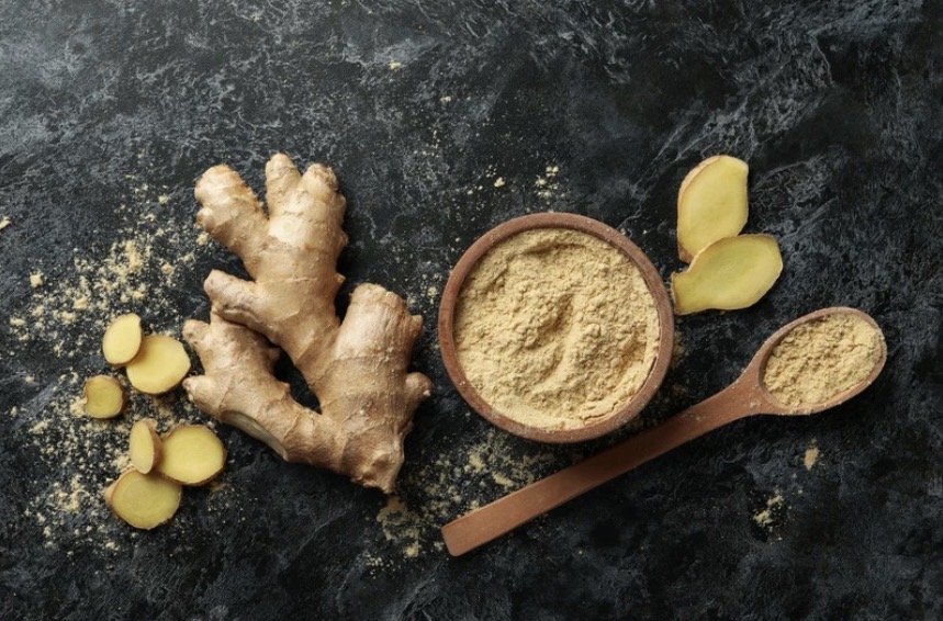 Fresh ginger root, sliced coins, and ground ginger powder on a dark surface.