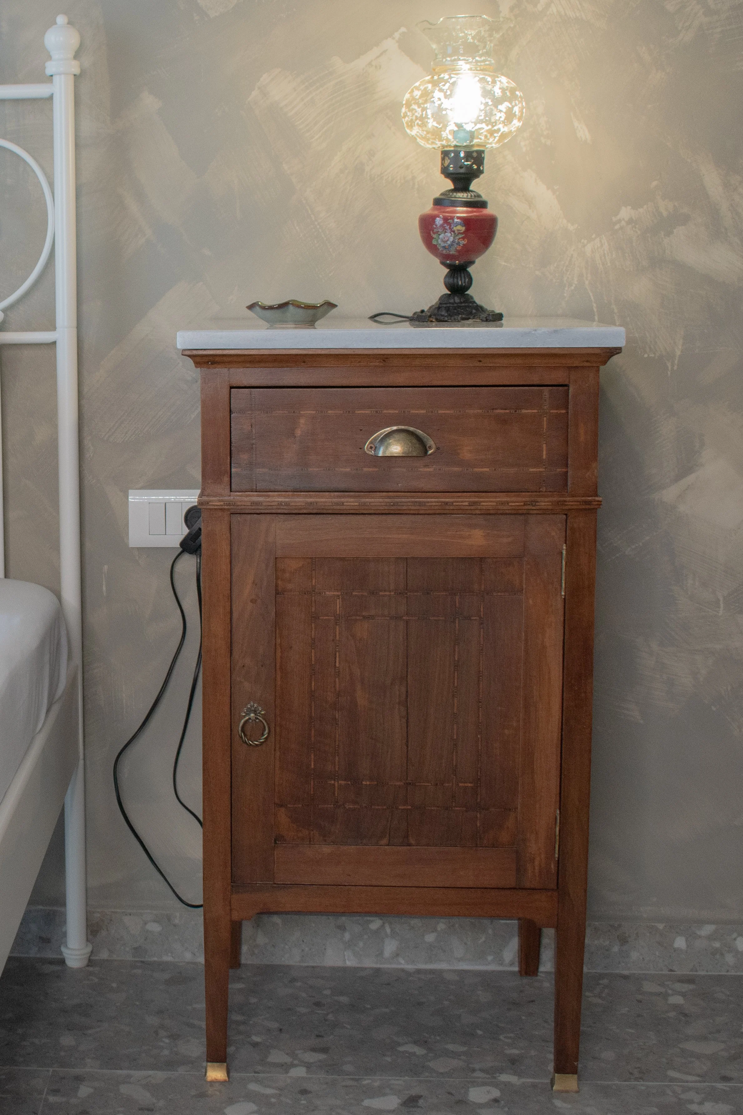 Wooden nightstand with drawer and cabinet, decorative table lamp, ceramic dish, power outlet, marble floor, textured wall background.