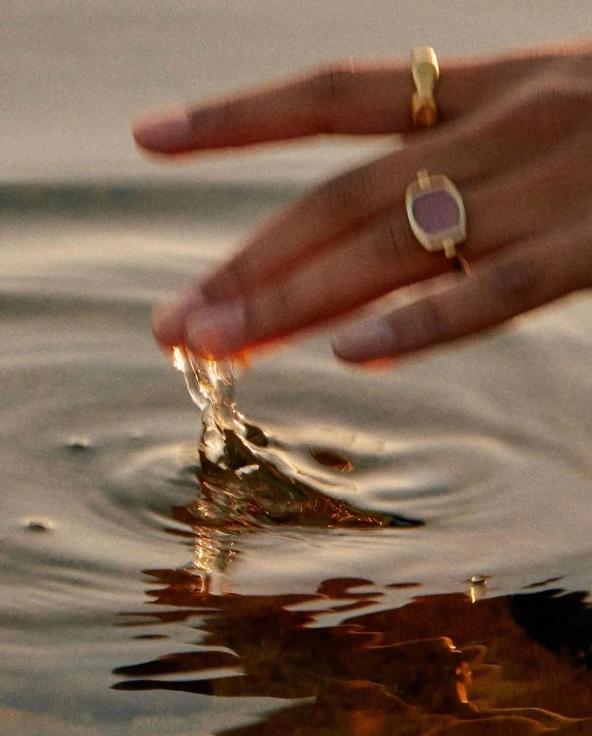 Nahaufnahme einer Hand, die im Wasser spielt, mit goldenen Ringen und einem Lavendel-Farbigen Nagellack. Dabei entstehen Wasserbewegungen und Tropfen.