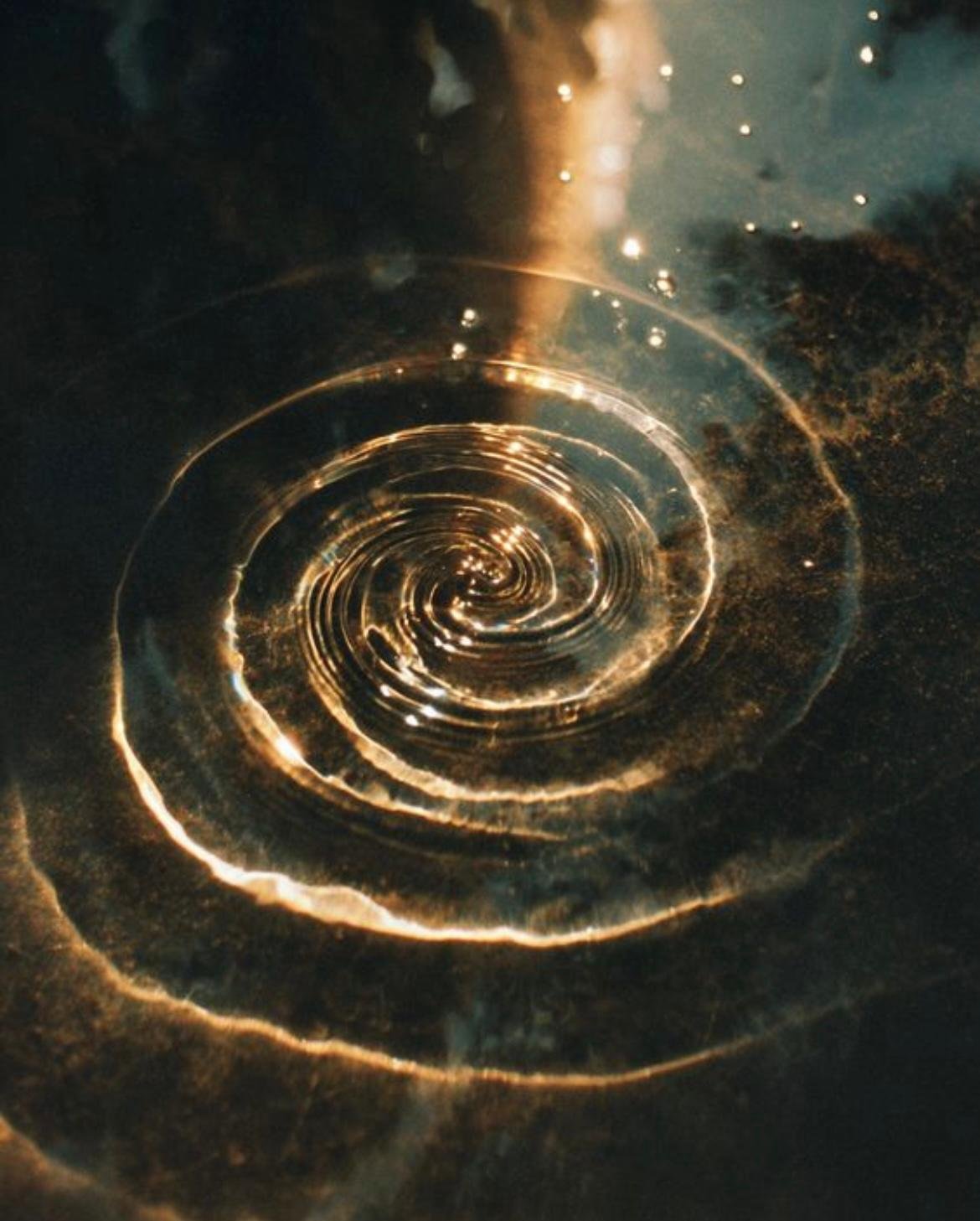 Nahaufnahme einer spiralförmigen Wasserlache auf einer dunklen Oberfläche, das Licht reflektiert auf der Wasseroberfläche.