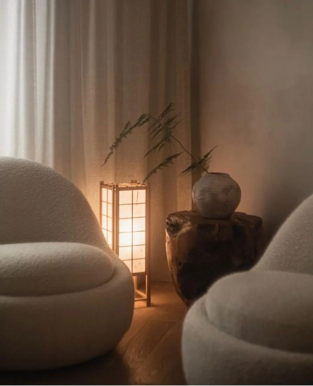 Gemütliches Wohnzimmer mit zwei weißen Sesseln, einem Holztisch, einem Kerzenständer mit Lampe und einem Vase mit Zweigen.