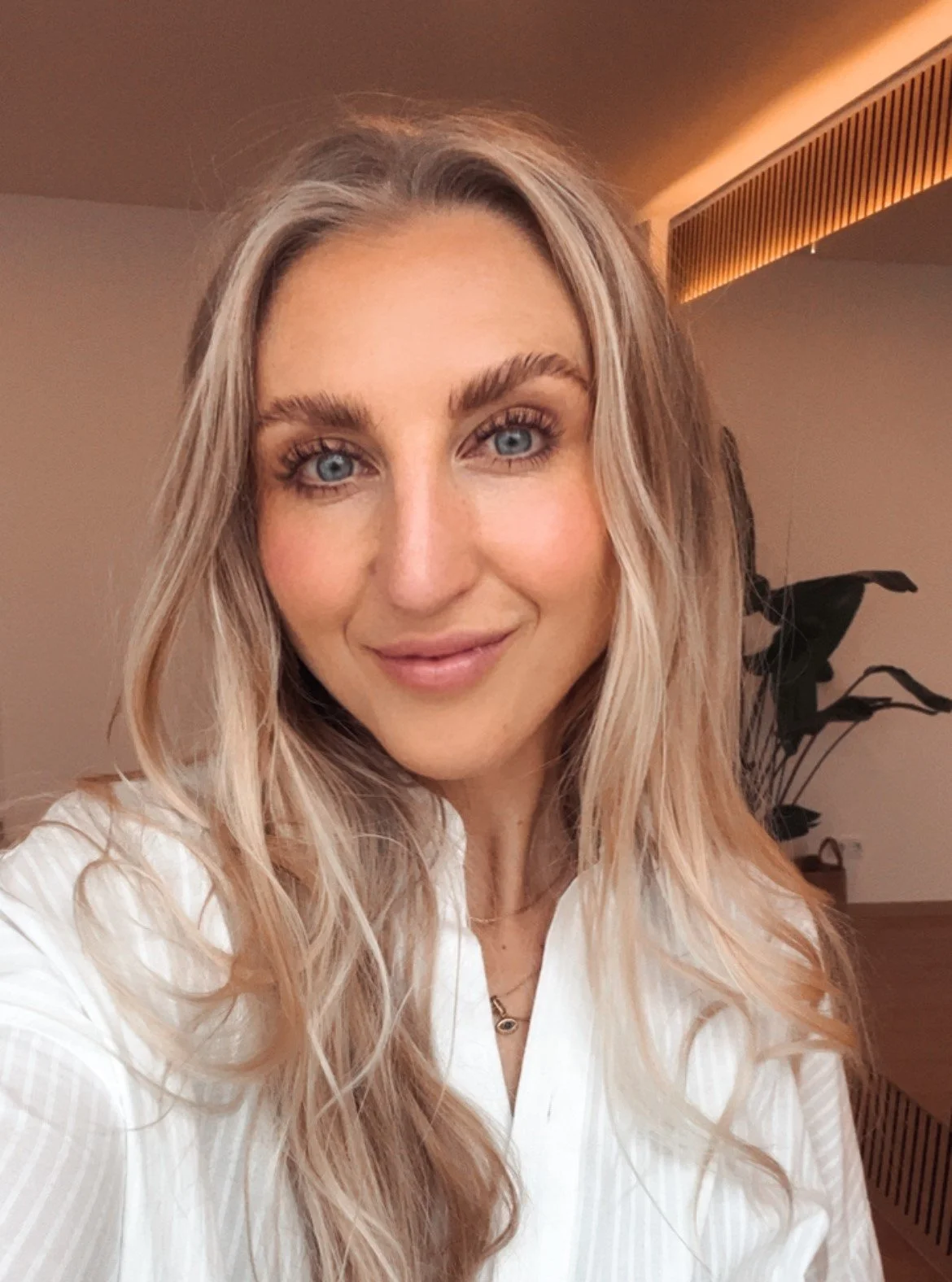 Ein Porträt einer jungen Frau mit blonden, gewellten Haaren und blauen Augen. Sie trägt ein weißes Hemd und hat ein freundliches Lächeln. Im Hintergrund sind eine gedeckte Wand, ein großes Pflanzblatt und eine indirekte Beleuchtung sichtbar.