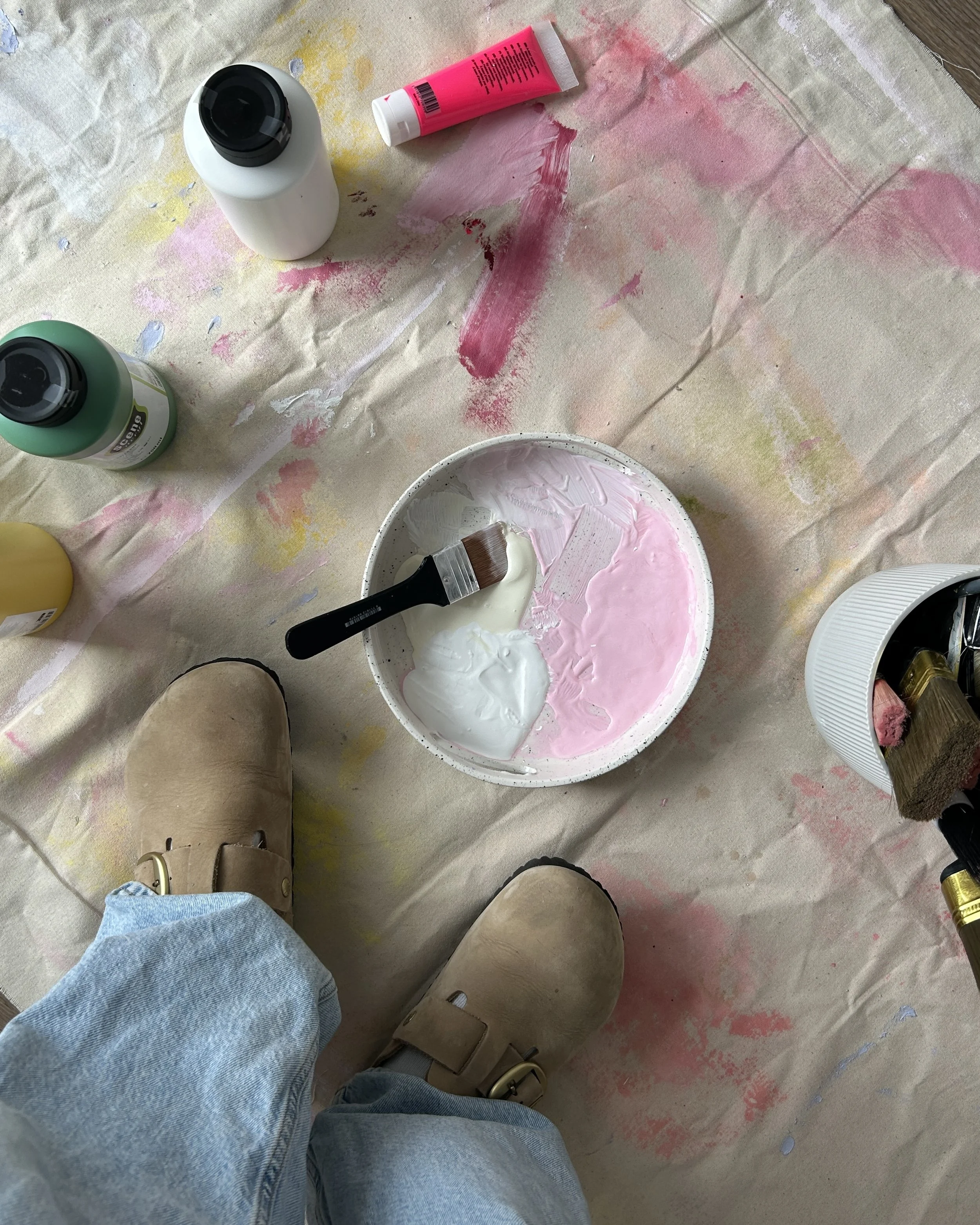 Fußboden mit bemalten Farben und Pinsel, umgeben von Farbflaschen und einem Schüssel mit weißer und rosa Farbe, Schuhen und Jeans.