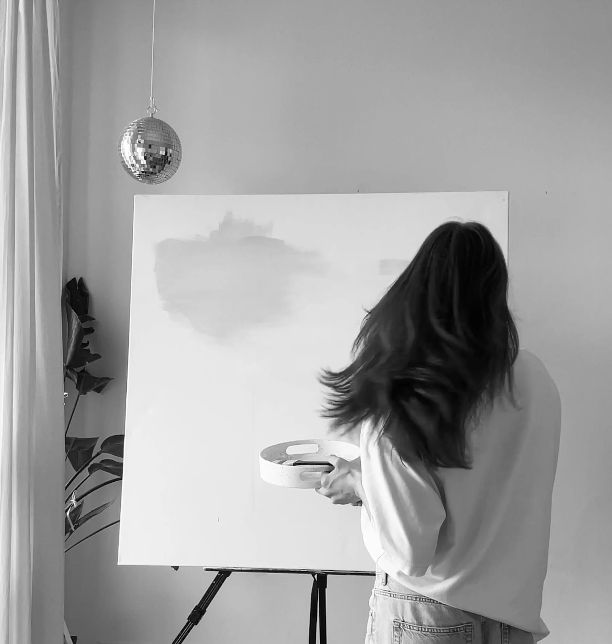 Eine Frau mit langen Haaren, die eine Palette mit Farben hält, steht vor einer leeren Leinwand und malt. Es hängt ein Disco-Kugel an der Decke, und im Hintergrund sind Pflanzen zu sehen.