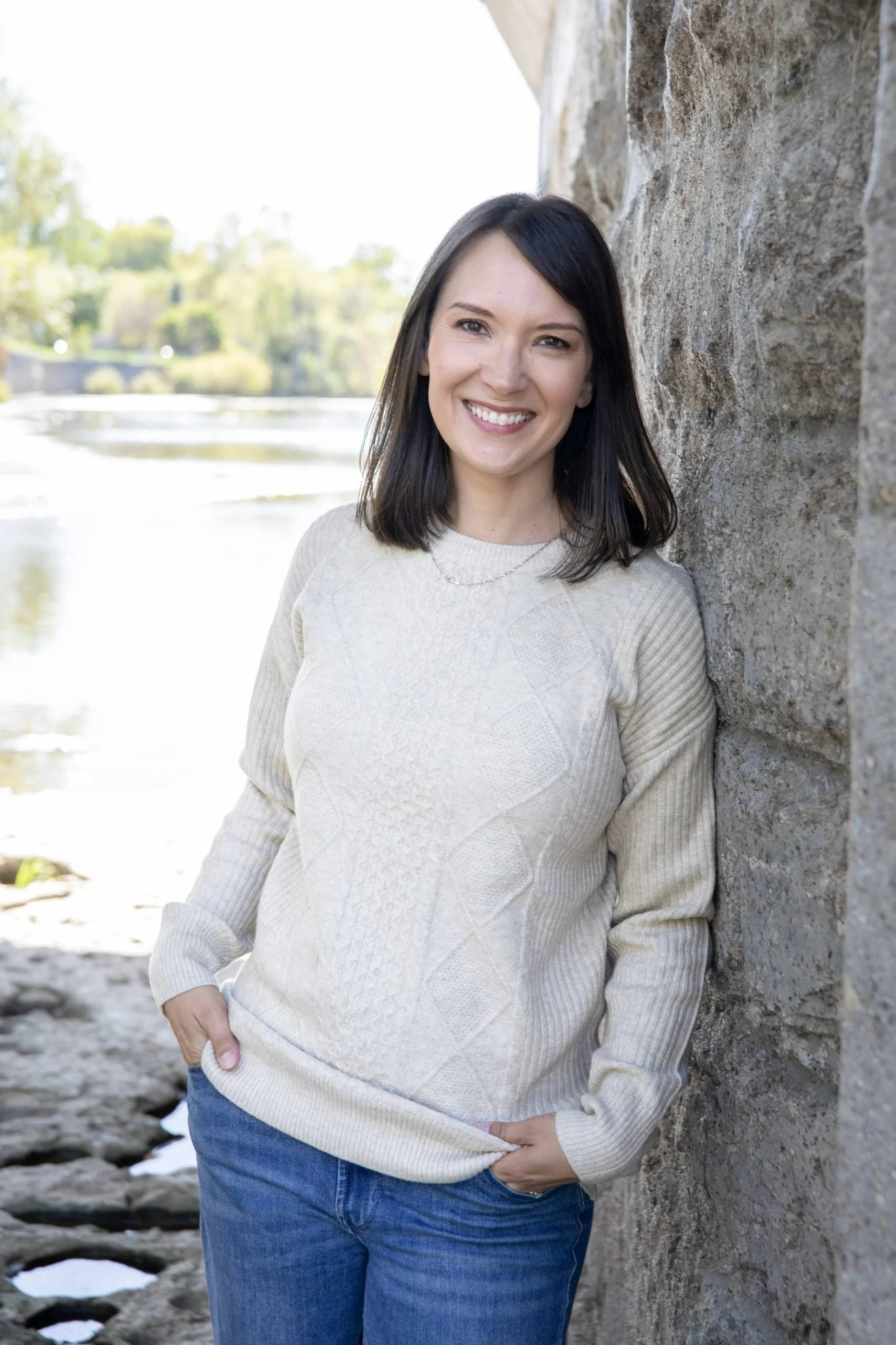A woman with shoulder-length dark hair smiling, wearing a beige sweater and jeans, leaning against a stone wall outdoors near a body of water on a sunny day.