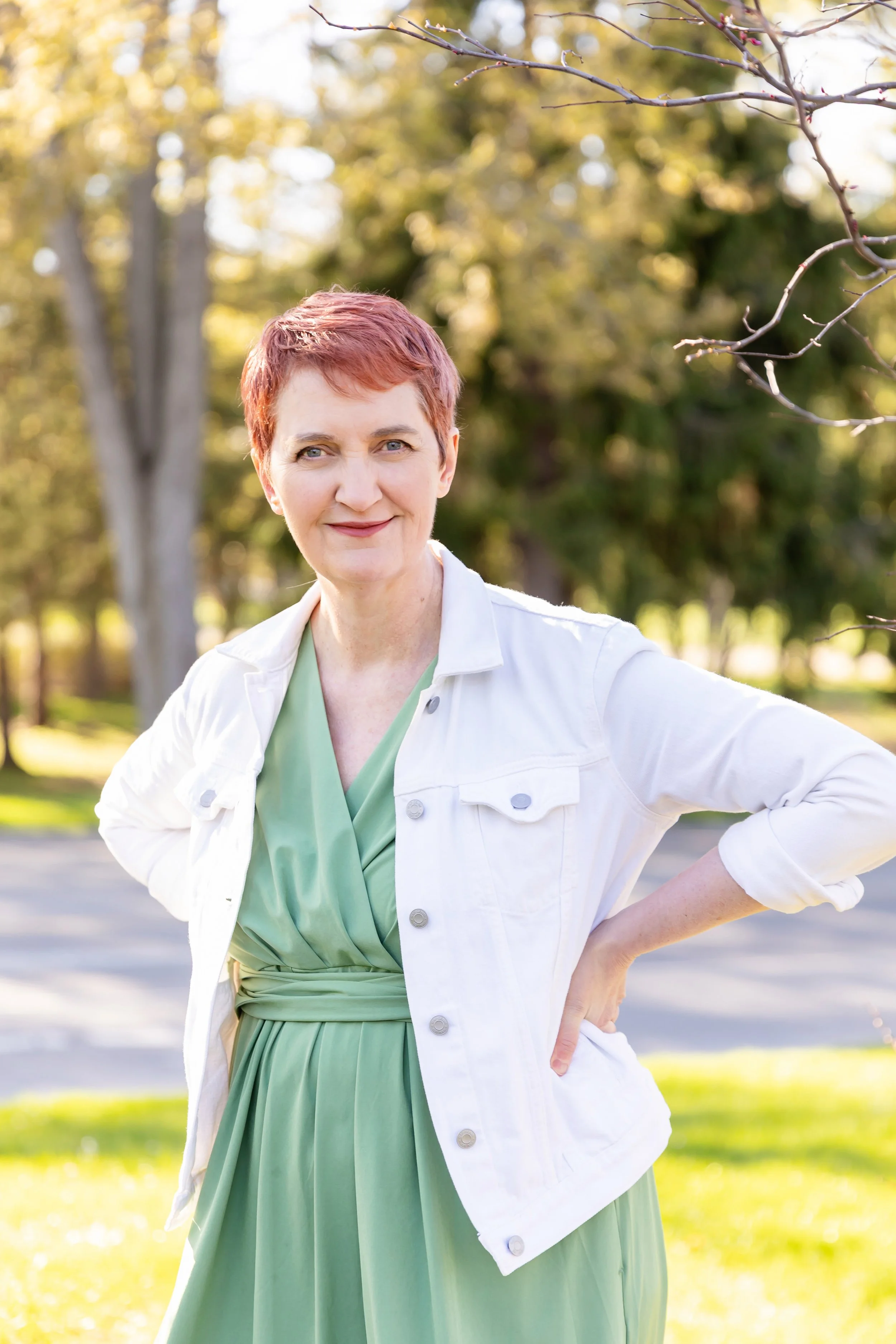 A woman with short reddish hair smiling outdoors in a park, wearing a light green dress and a white jacket with sleeves rolled up, with trees and sunlight in the background.