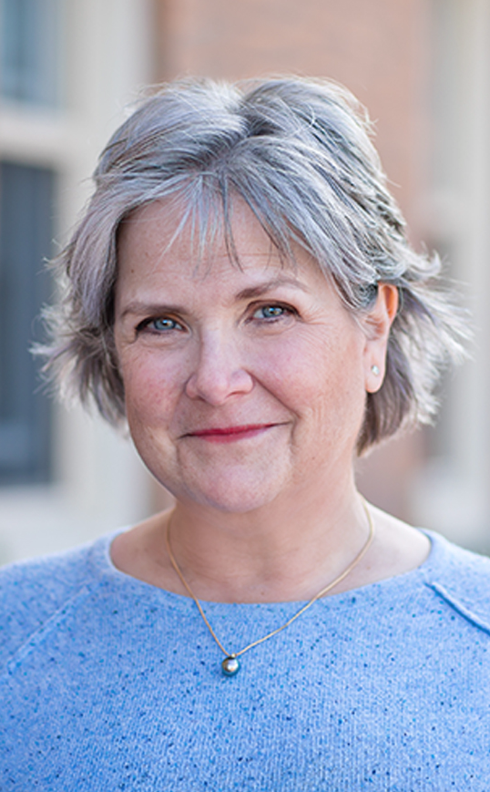 A portrait of a middle-aged woman with short gray hair, blue eyes, and light skin, wearing a blue sweater and a gold necklace with a pearl pendant, standing outdoors with a blurred background of buildings.