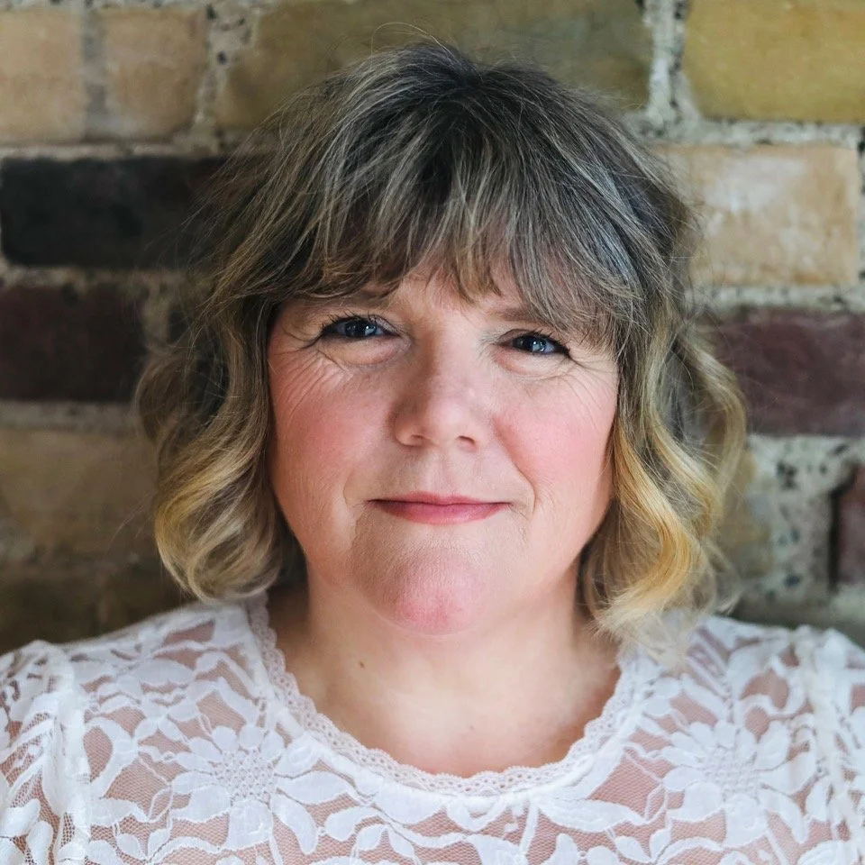 Close-up portrait of a woman with wavy blonde hair and blue eyes, wearing a white lace top, against a brick wall background.