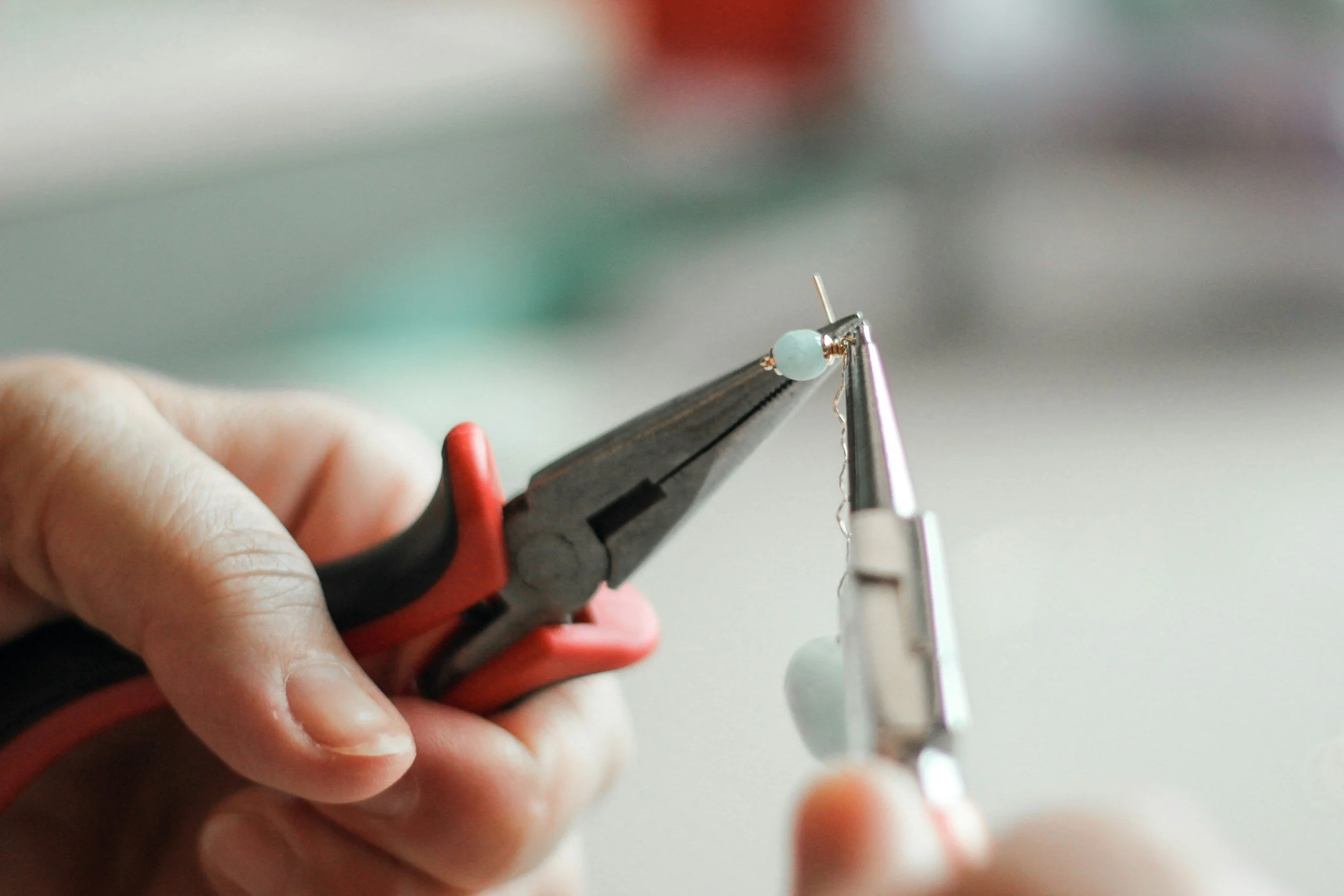 Close-up of hands holding jewelry pliers and tweezers, working on a small light blue bead, in a jewelry-making process.