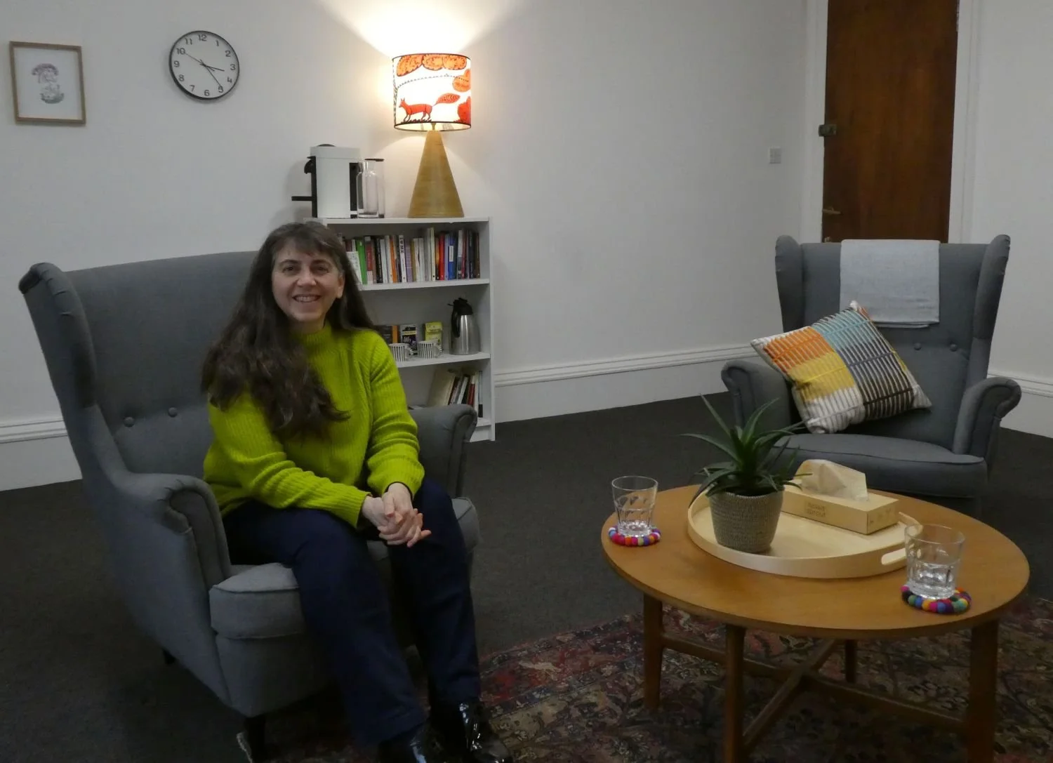 Counsellor seated in a comfortable counselling room with armchairs, bookcase, lamp and a coffee table with a plant, box of tissues and glasses of water on it