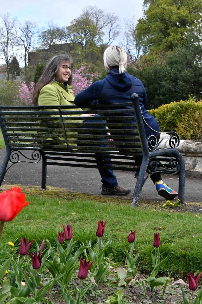 Helen sitting on a park bench with a client in the spring with flowers in the foreground and trees in blossom in the background