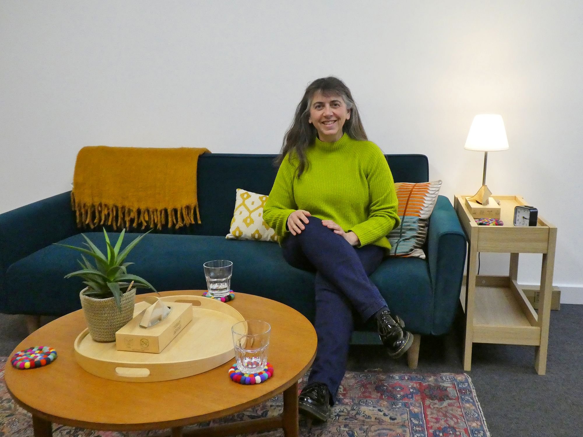 Helen seated on a sofa with cushions in a comfortable counselling room with a table in the foreground with a plant, box of tissues and glasses of water on it