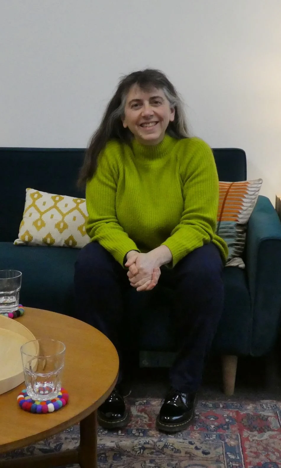 Helen sitting on a sofa with cushions in a counselling room next to a coffee table with glasses of water