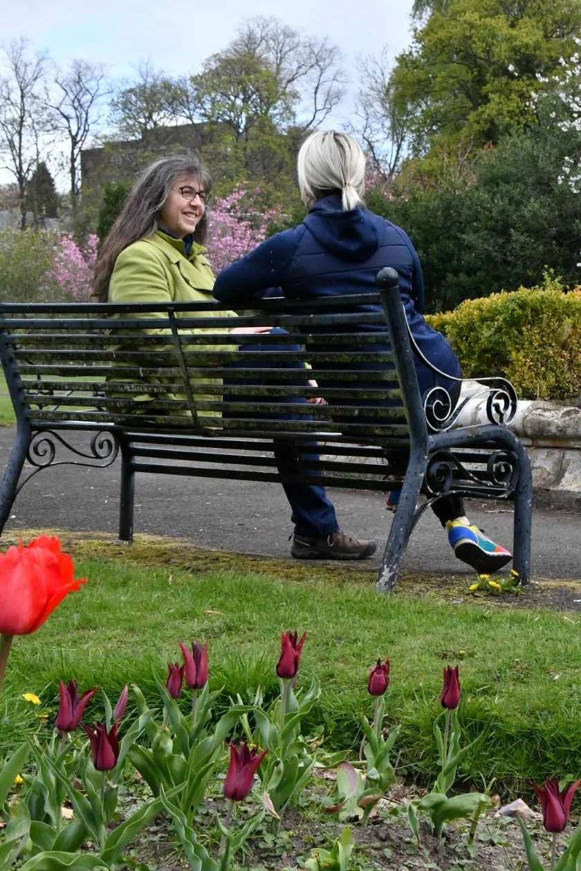 Helen sitting on a park bench with a client in the spring with trees in blossom in the background