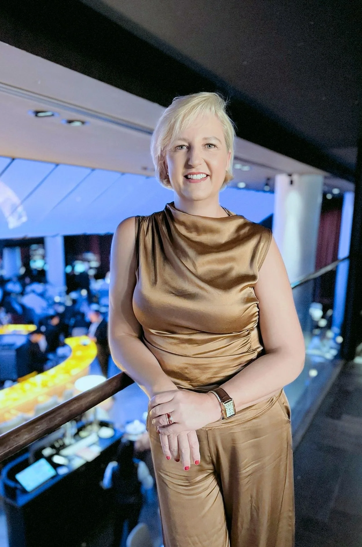 A smiling woman with blonde, chin-length hair, wearing pearl earrings and a blue scarf, outdoors with blurred background lights.