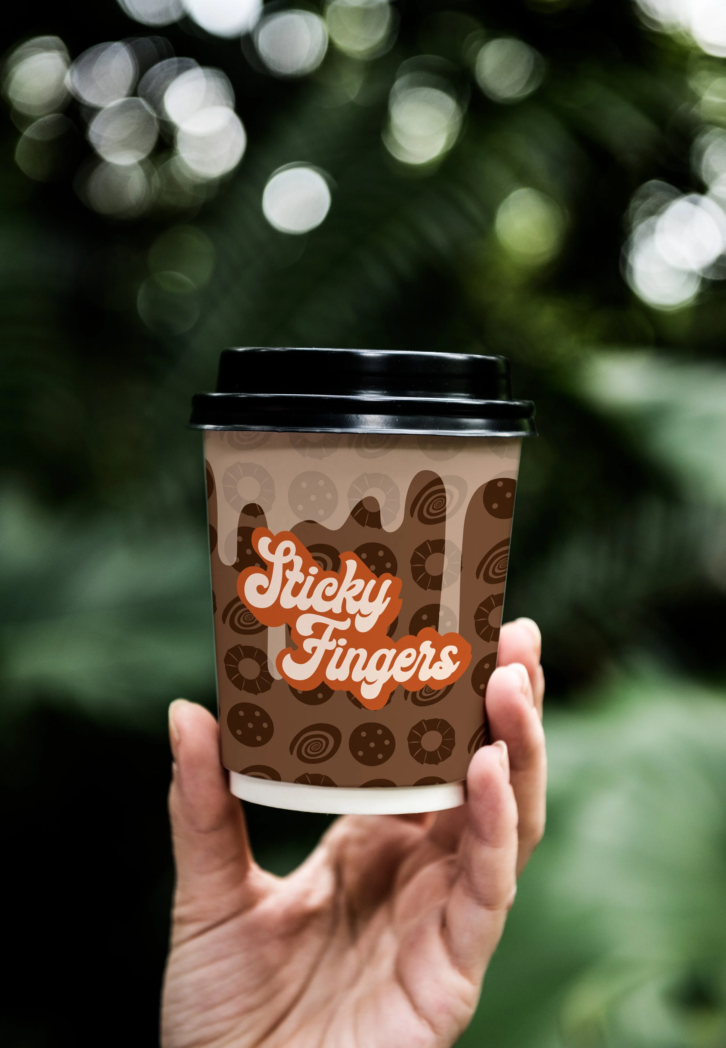 Hand holding a paper coffee cup with a black lid, labeled 'Sticky Fingers,' with a background of green foliage.