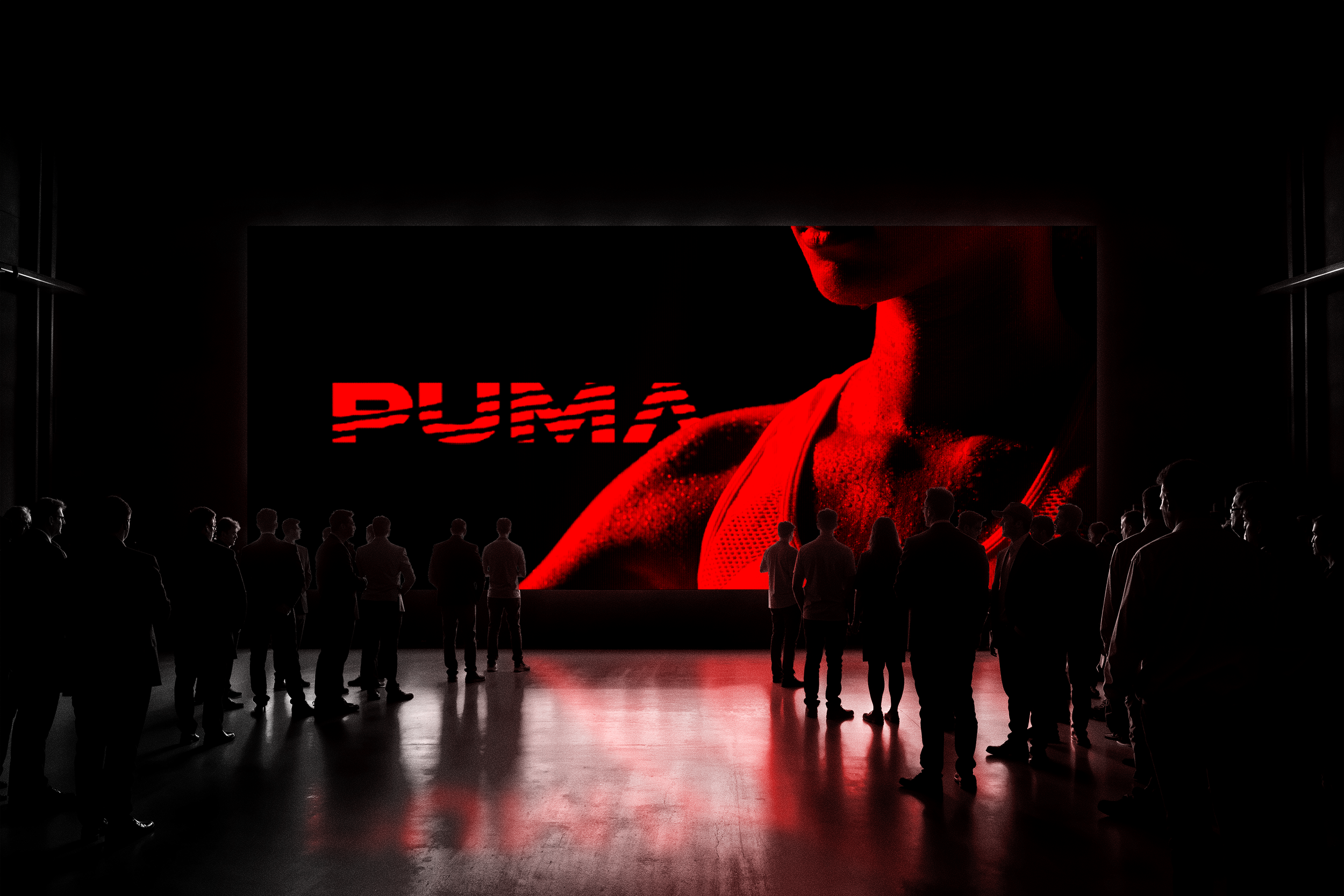 Group of people watching a large screen with a red image of a woman's face and the word 'PUMA' on a black background in a dimly lit room.