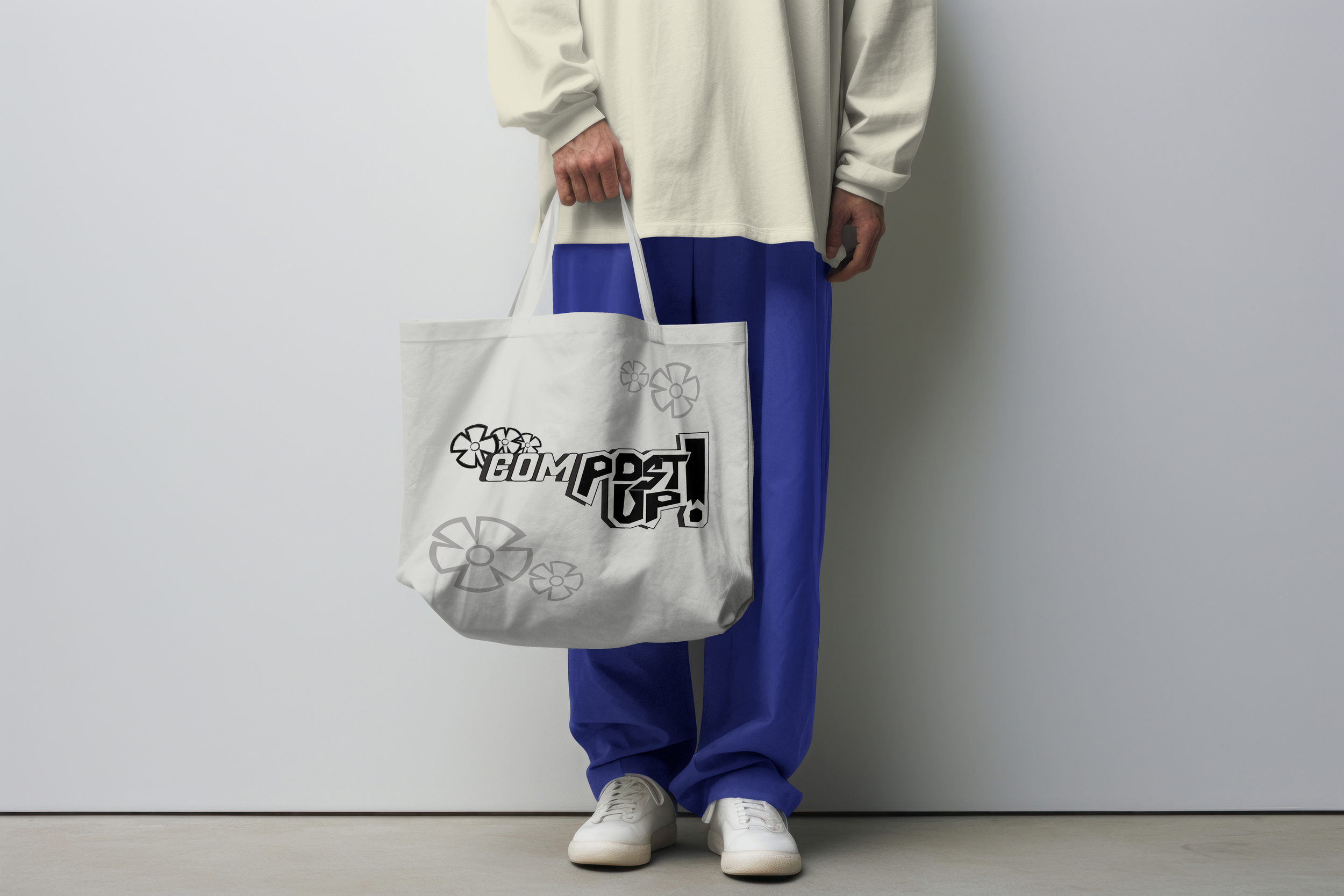 Person holding a white tote bag with a black and white graphic design that reads 'COMPOST UP!' and flower illustrations, wearing a beige long-sleeve top, blue pants, and white sneakers, standing against a plain white wall.