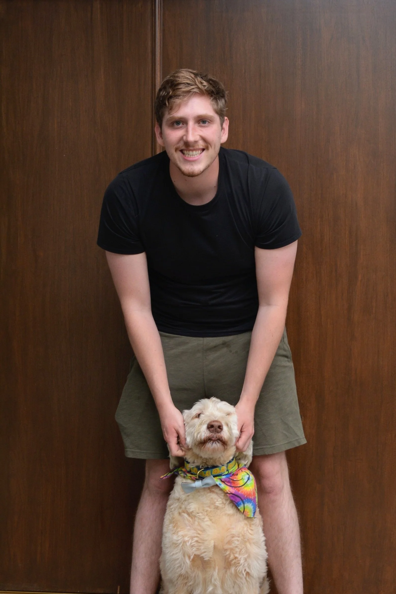 A young man with light brown hair, wearing a black t-shirt and shorts, smiling while holding the dog's head from behind. The dog is a golden retriever with curly fur, wearing a colorful bandana. They are in front of a wooden wall.