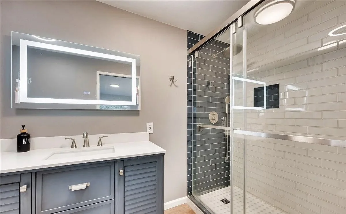 Modern bathroom featuring a blue vanity with a white countertop and a large illuminated mirror, a black soap dispenser, and a walk-in shower with glass doors, dark gray tiles, and a white-tiled shower wall.