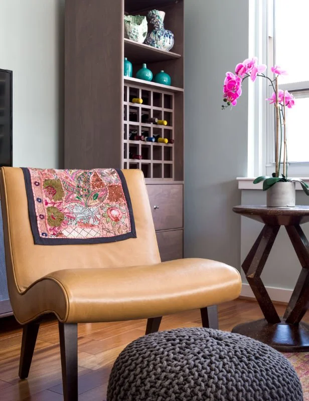 A cozy corner with a tan leather chair, a colorful embroidered pillow, a knitted gray pouf on a wooden floor, a side table with a pink orchid, and a tall wooden bookshelf with decorative items and a wine rack near a window.