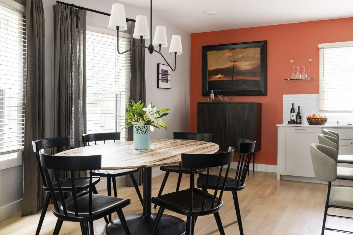Dining room with a round wooden table, six black chairs, a centerpiece with white lilies in a turquoise pot, large windows with blinds and dark curtains, a modern chandelier, artwork on the wall, a dark cabinet, a framed painting, a white sideboard w