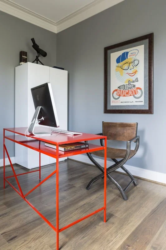 A modern home office with a red metal desk holding an iMac computer, a vintage wooden chair, a white cabinet with a telescope and a book, and a framed vintage motorcycle poster on a gray wall.