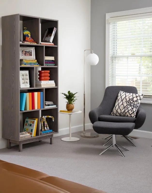 A cozy reading nook with a tall gray bookshelf filled with colorful books, a small white side table with a potted plant, a gray armchair with a geometric pillow, a standing white floor lamp, and a window with blinds letting in natural light.