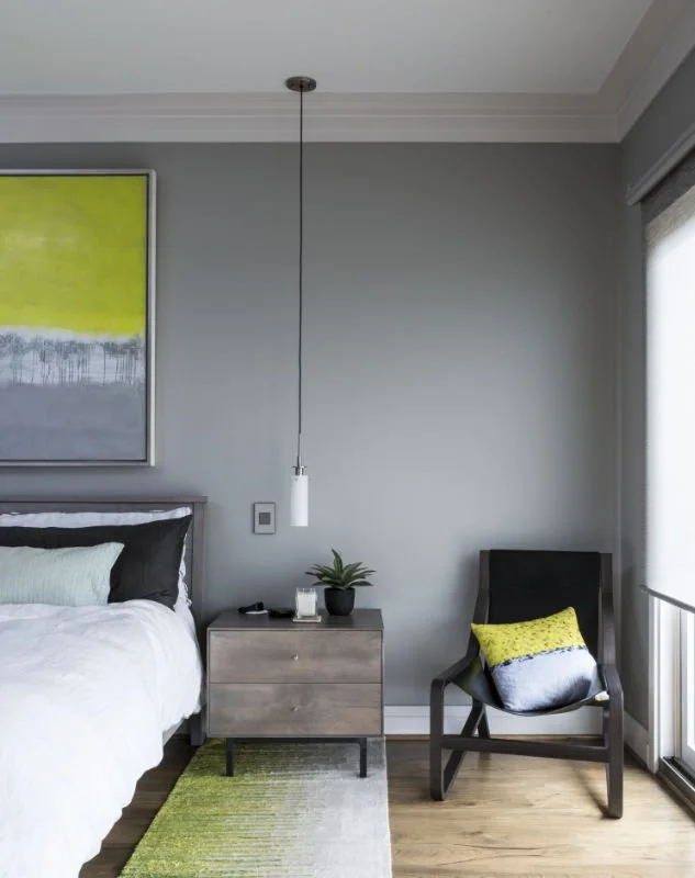 A modern bedroom with gray walls, a bed with white and black bedding, a wooden nightstand with a plant, candle, and remote, a black armchair with a colorful pillow, and abstract artwork with yellow and gray tones on the wall.