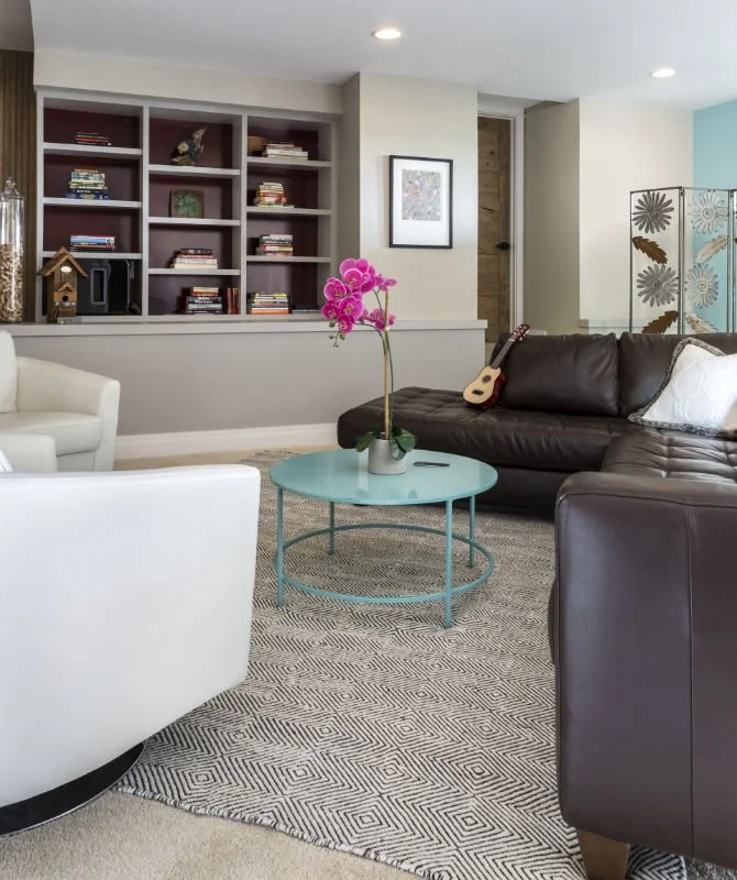 Living room with a dark brown leather sofa, a white armchair, a glowing orchid plant on a pastel blue coffee table, a bookshelf with books, a framed picture, a guitar, and a decorative folding screen with floral patterns.