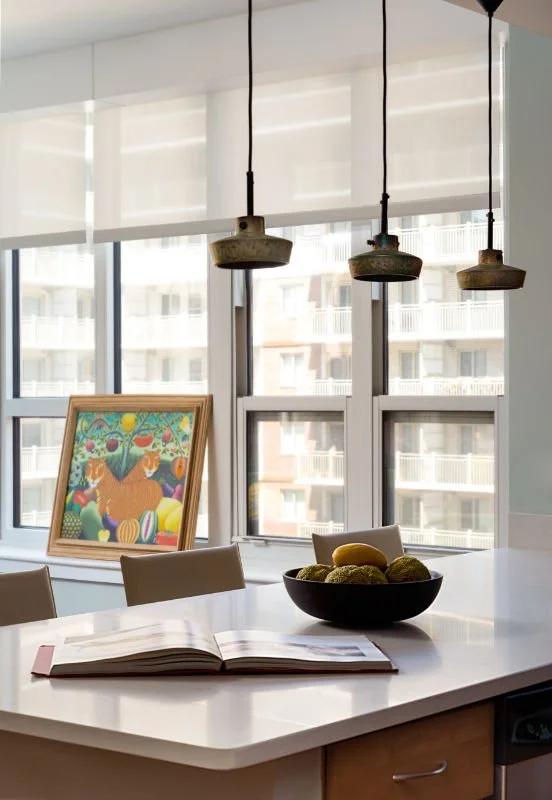Bright kitchen with large windows, hanging pendant lights, a bowl of lemons and limes, an open book on a white countertop, and a colorful framed artwork of a tiger surrounded by fruits.