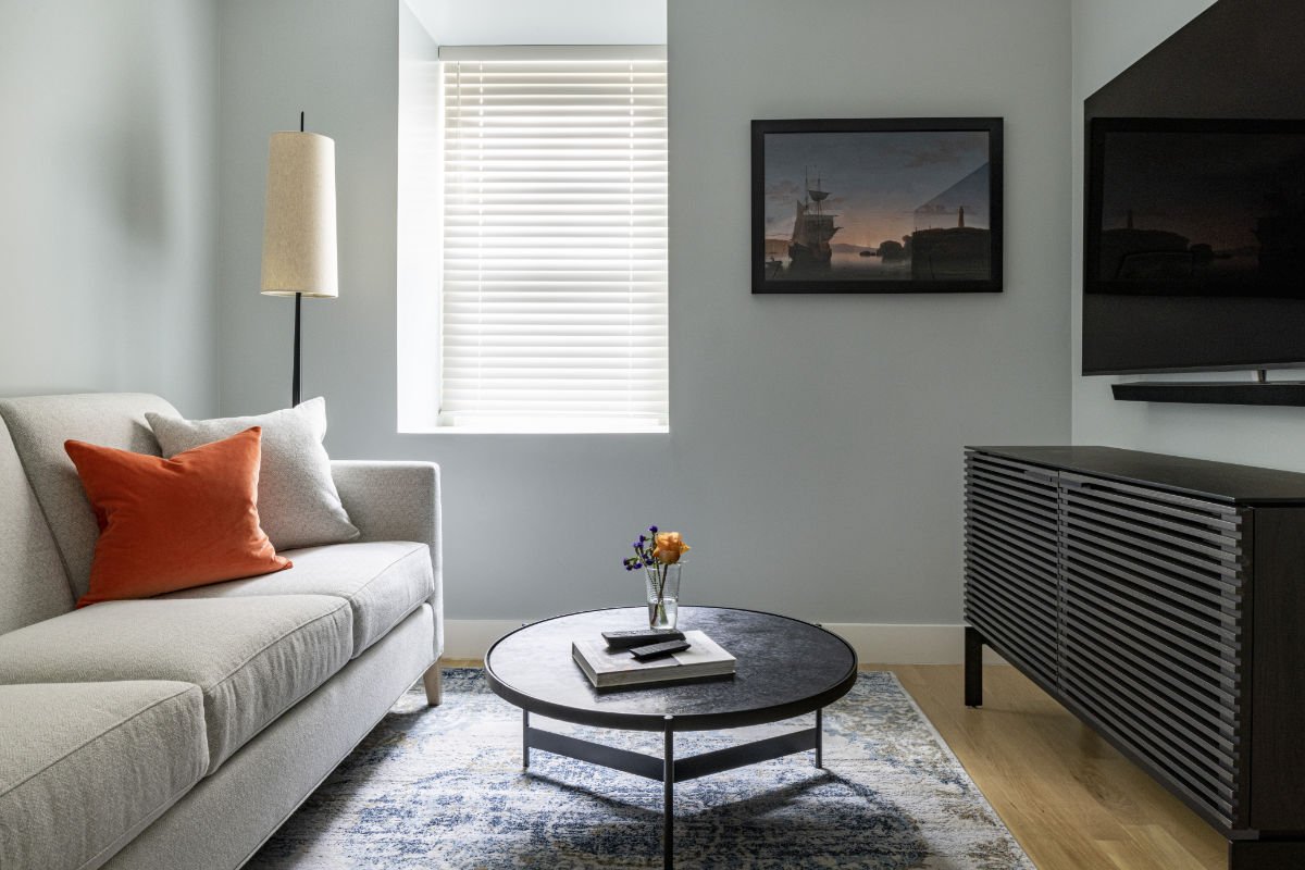 Living room with a beige sofa, orange and light gray pillows, a tall floor lamp, a window with closed blinds, a wall art of ships at sunset, a black round coffee table with a vase and books, a patterned rug, and a black TV stand with a flat-screen TV