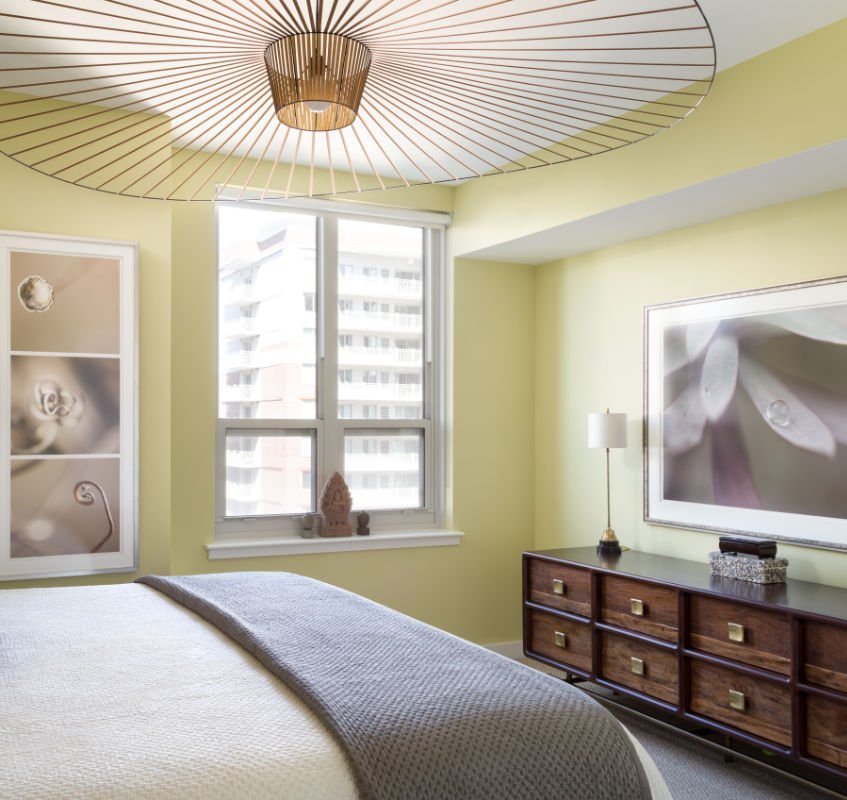 A bedroom with a bed covered in a textured gray blanket, a window with views of a high-rise building, a wooden dresser with gold handles, a tall white lamp, art on the walls including a large framed flower and a bonsai sculpture on the windowsill, an