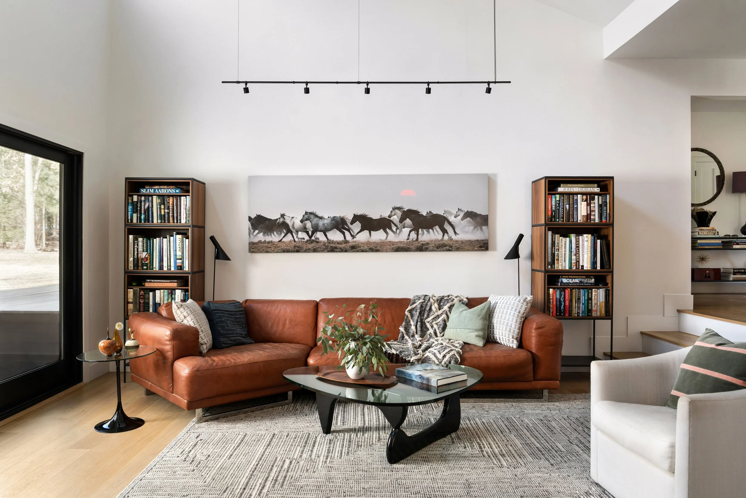 Living room with a brown leather sofa, white armchair, glass coffee table, bookshelves, wall art of running horses, and large sliding glass door with outdoor trees.