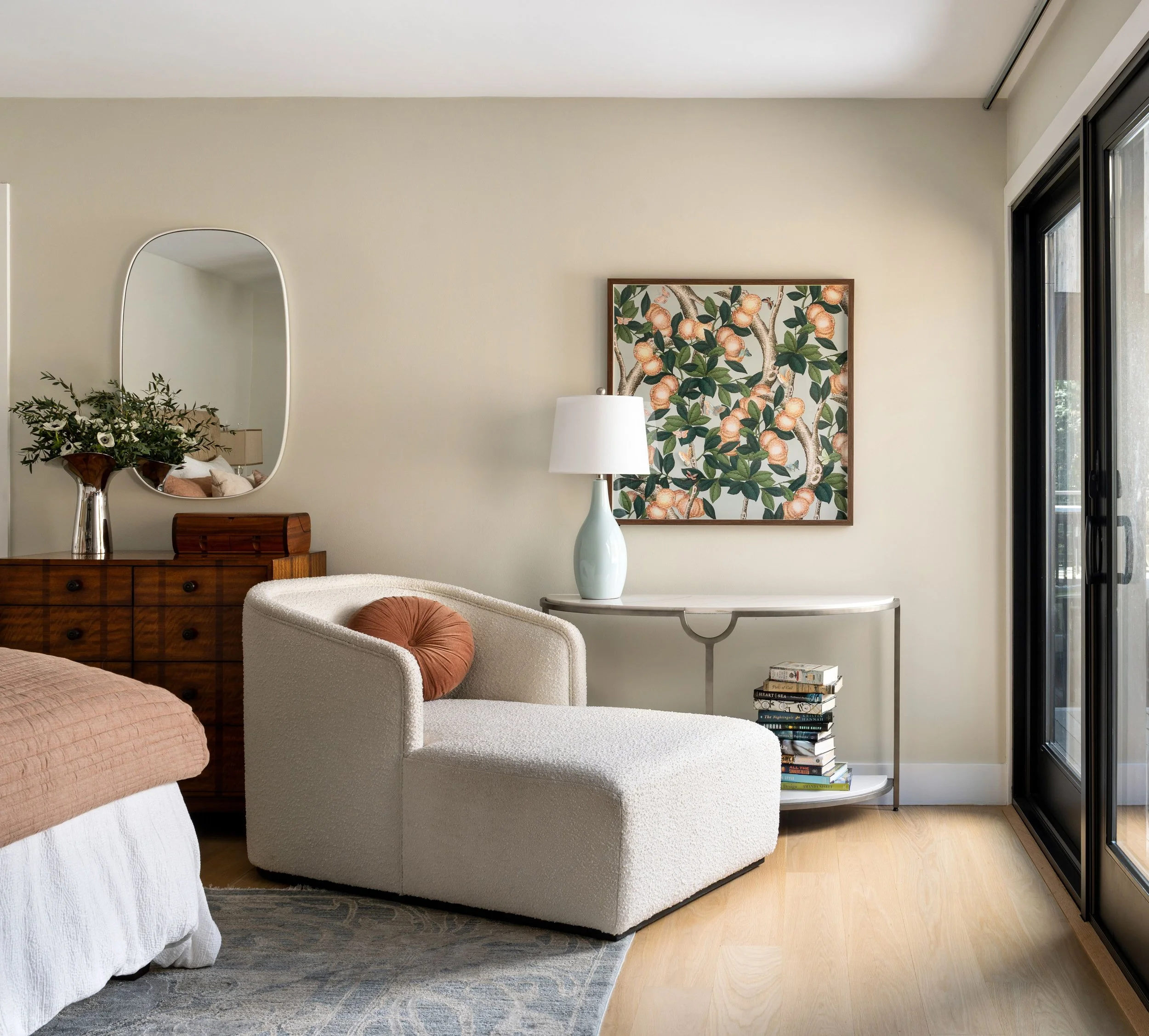 Living room corner with a white textured armchair, a wooden sideboard with a mirror and flowers, a white-console table with a lamp and stacked books, a framed botanical art on the wall, and a sliding glass door.