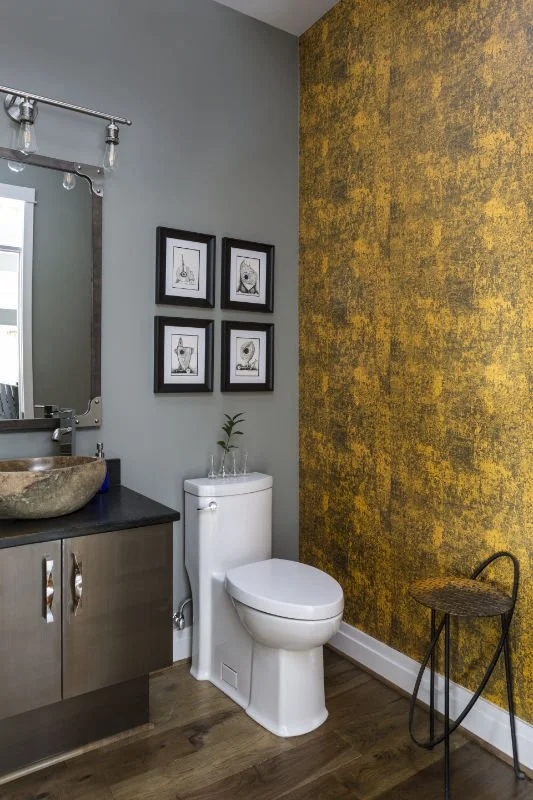 Small bathroom with a gray wall, a yellow textured accent wall, a white toilet, a vanity with a stone sink, a rectangular mirror, and four framed black-and-white artistic prints on the gray wall.