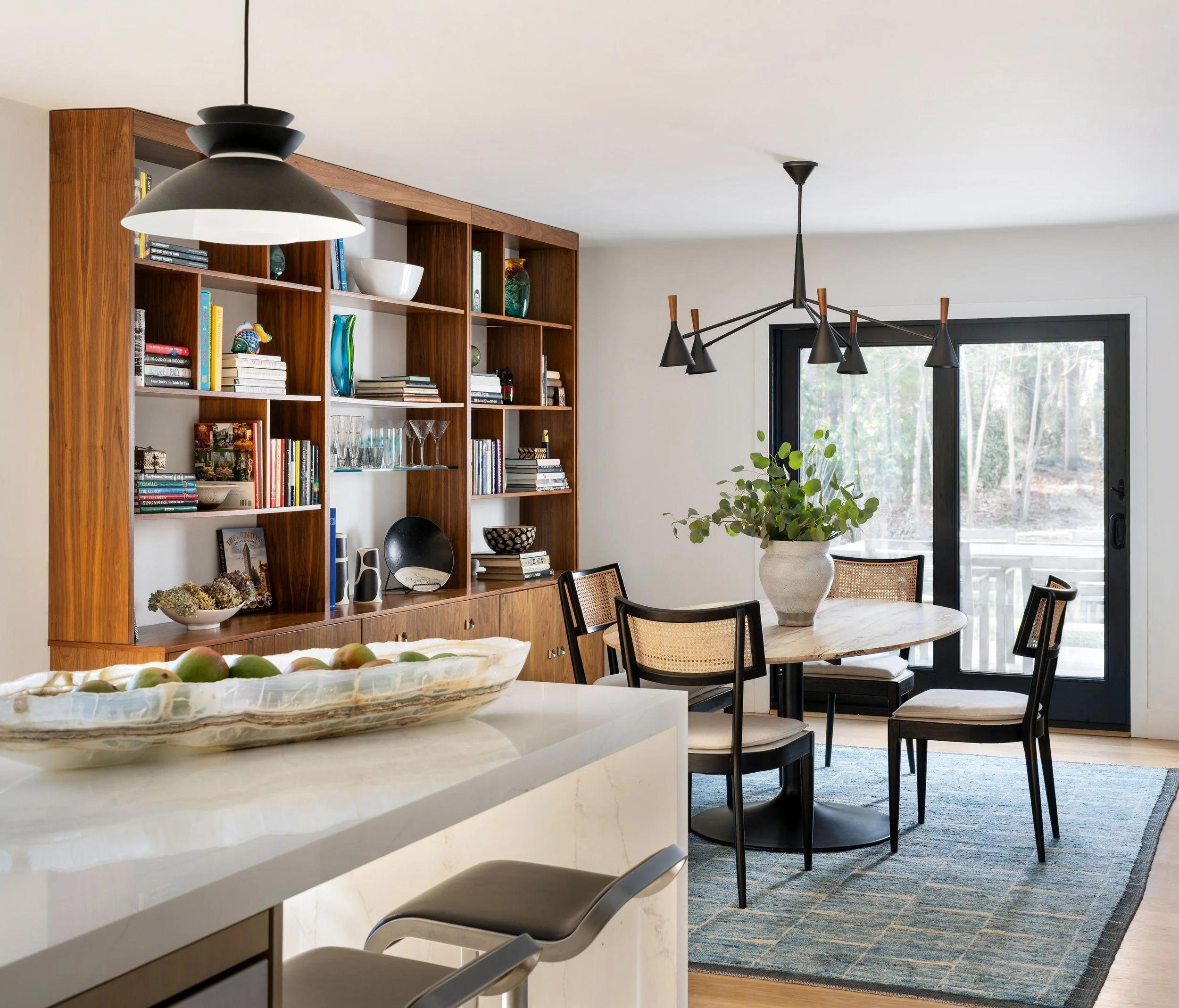 Modern dining room with a round table, four chairs, a large potted plant, a wooden bookshelf, and a sliding glass door leading to an outdoor area.