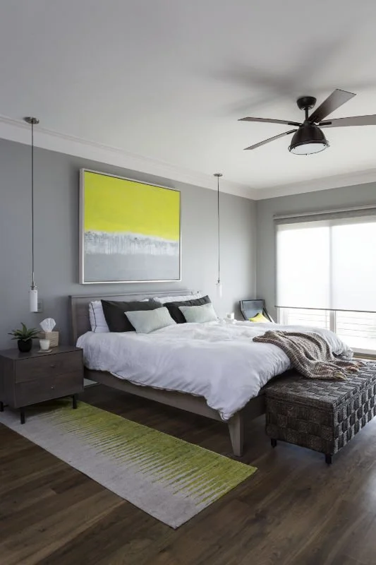 Modern bedroom with gray walls, a large bed with white linens and multiple pillows, a dark wooden nightstand with a plant and decor, a patterned ottoman at the bed's foot, abstract artwork with yellow and gray tones on the wall, hardwood floors, a ce