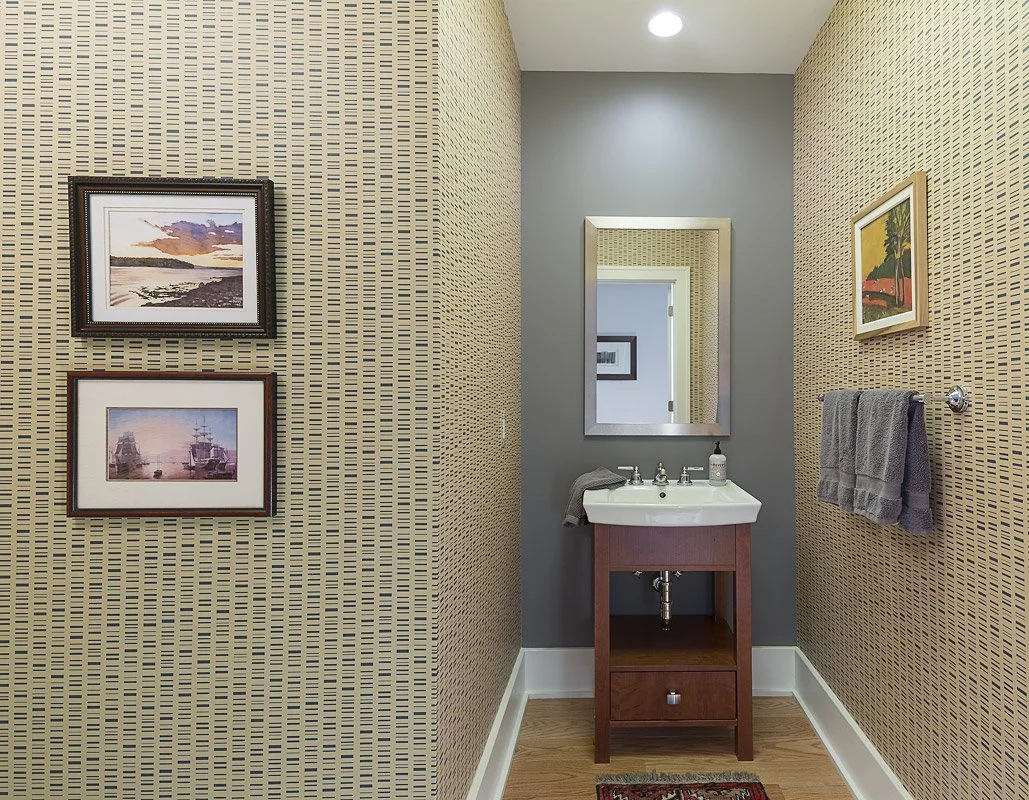 Small bathroom with patterned wallpaper, gray accent wall, a mirror above a small sink, framed art on the walls, and a gray towel hanging on a bar.